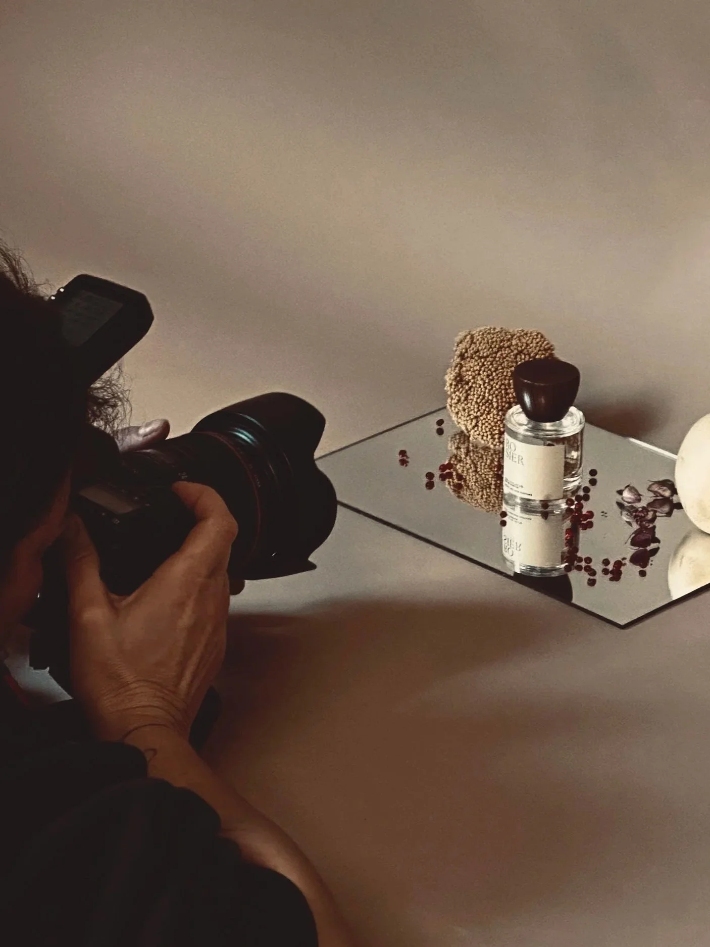 Photographer taking a photo of a perfume bottle, a piece of coral, and scattered red berries on a reflective surface.