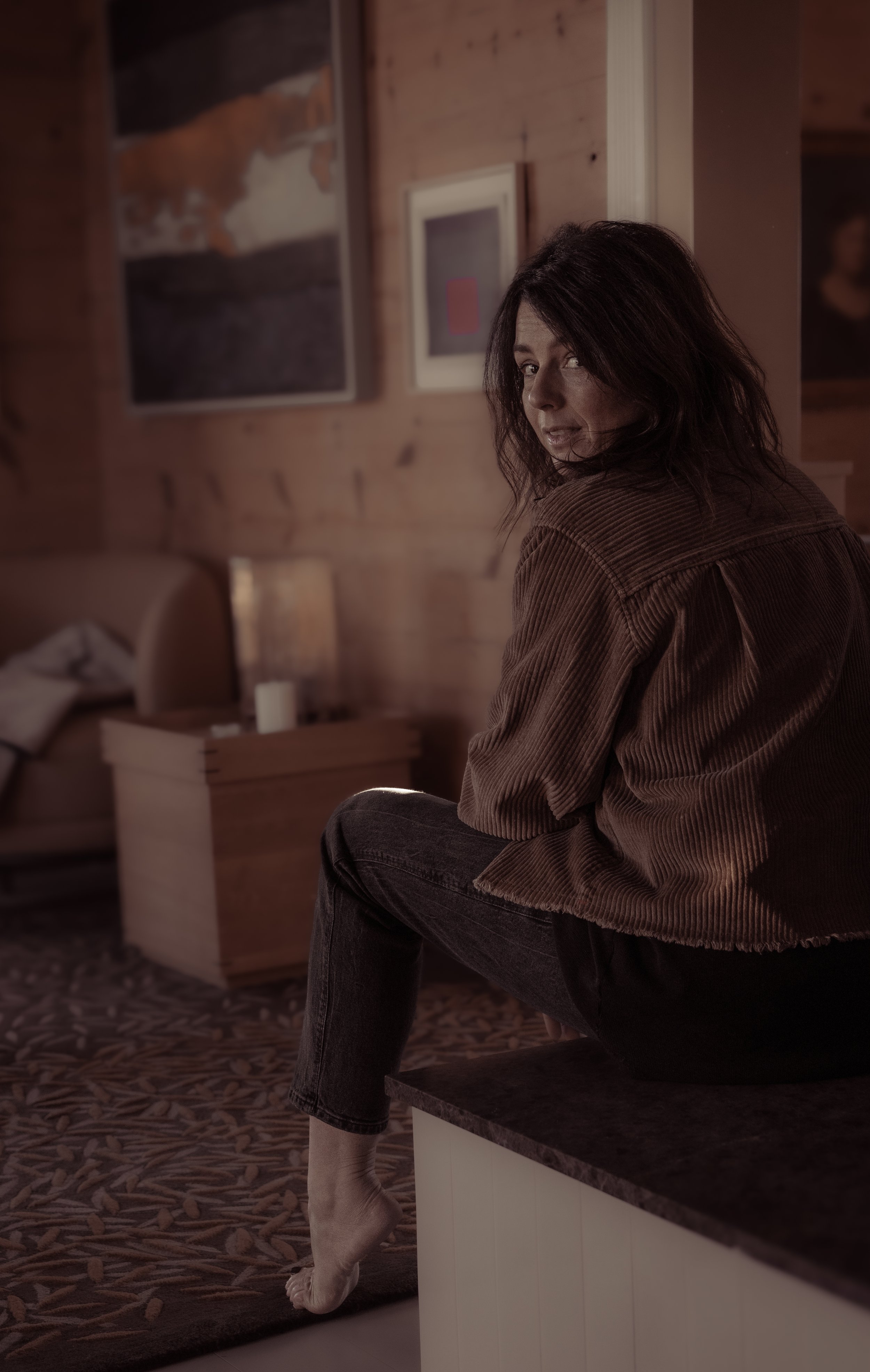 A woman sitting on a kitchen counter in a rustic, wood-paneled room, looking over her shoulder at the camera.