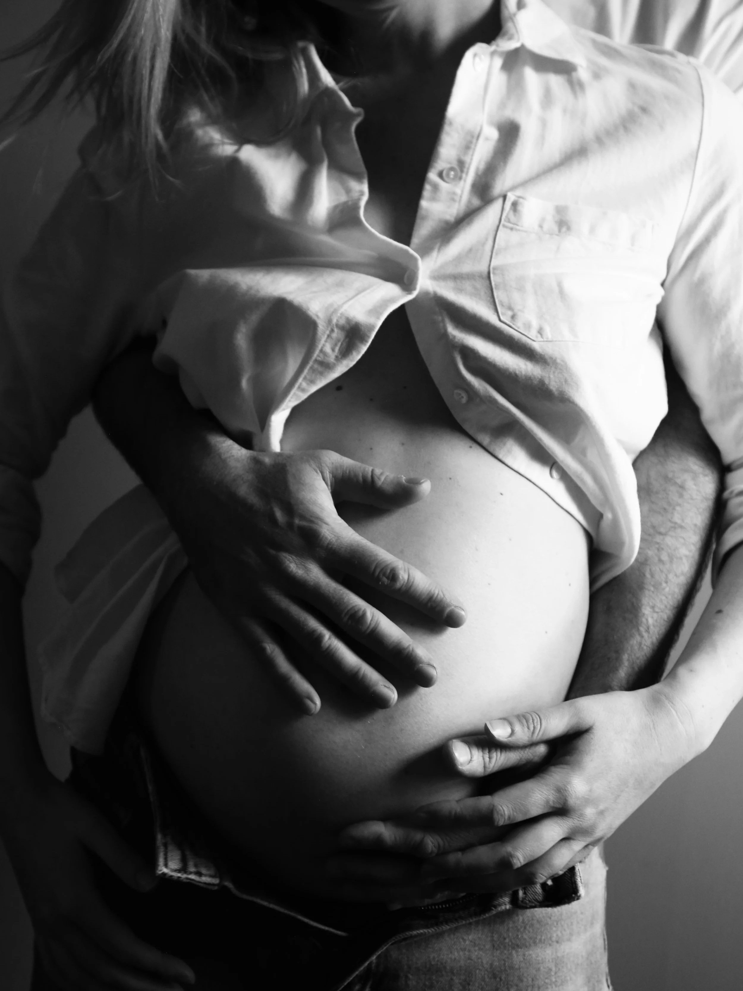 Close-up of a pregnant woman’s belly being held by her hands, with another person’s hands supporting from below, in a black and white photograph.