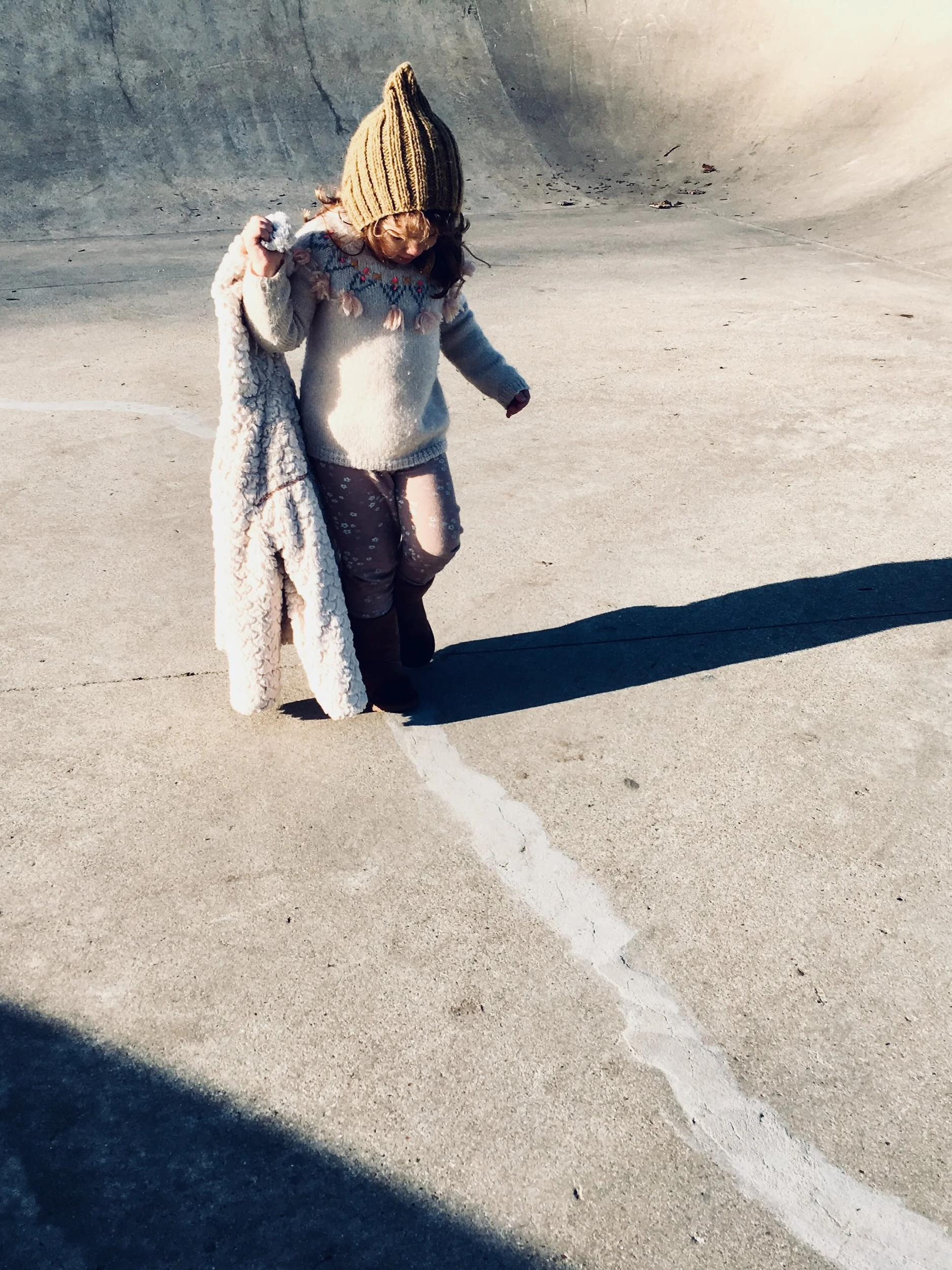 A young girl standing on a concrete skatepark border, wearing a beige knit hat, sweater, patterned pants, and boots, holding a large fluffy coat, with a long shadow cast on the ground.