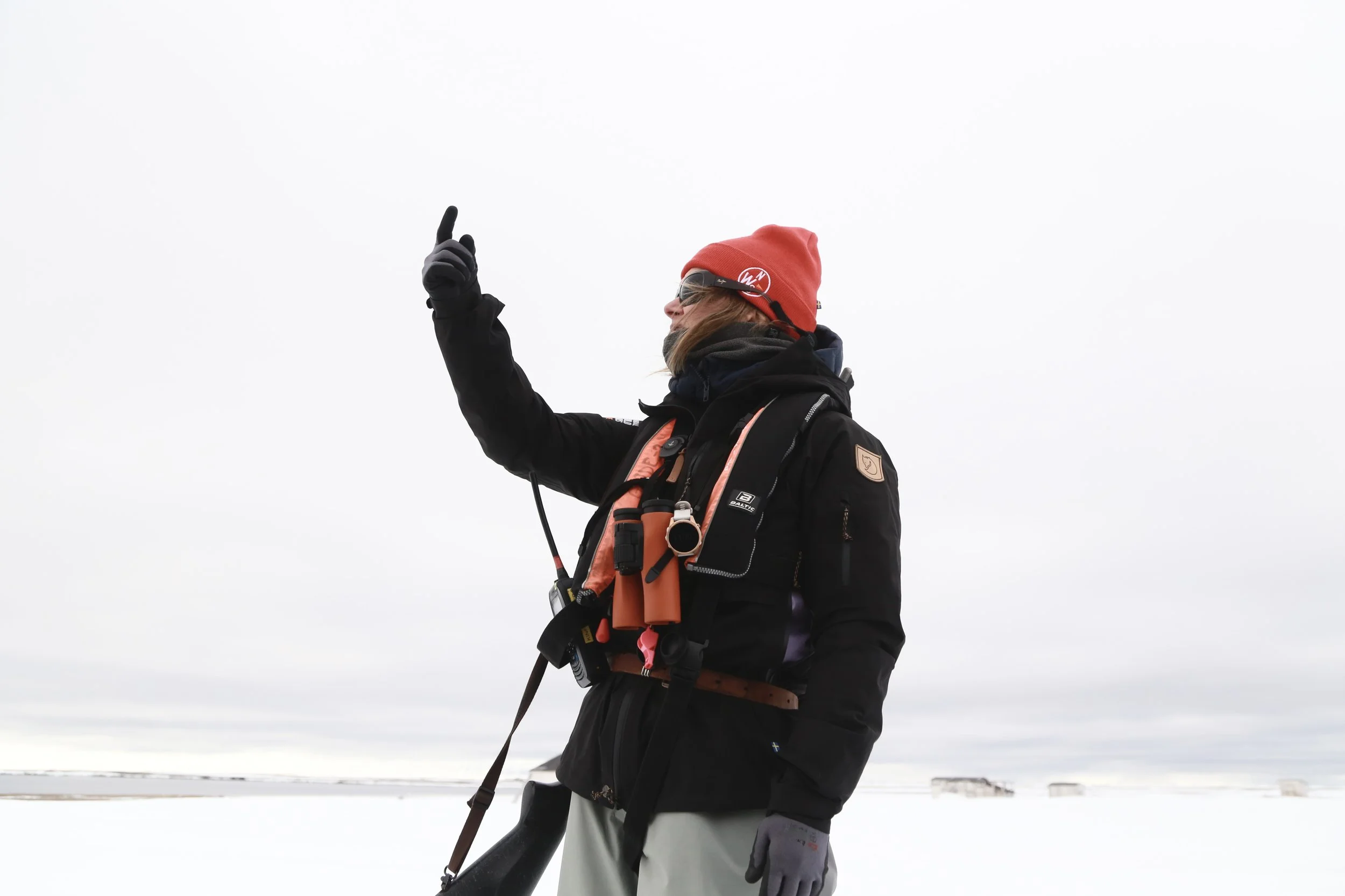 A person dressed in winter outdoor gear, wearing a red beanie, sunglasses, gloves, and a black jacket, standing in a snowy landscape and pointing upward with their right hand.