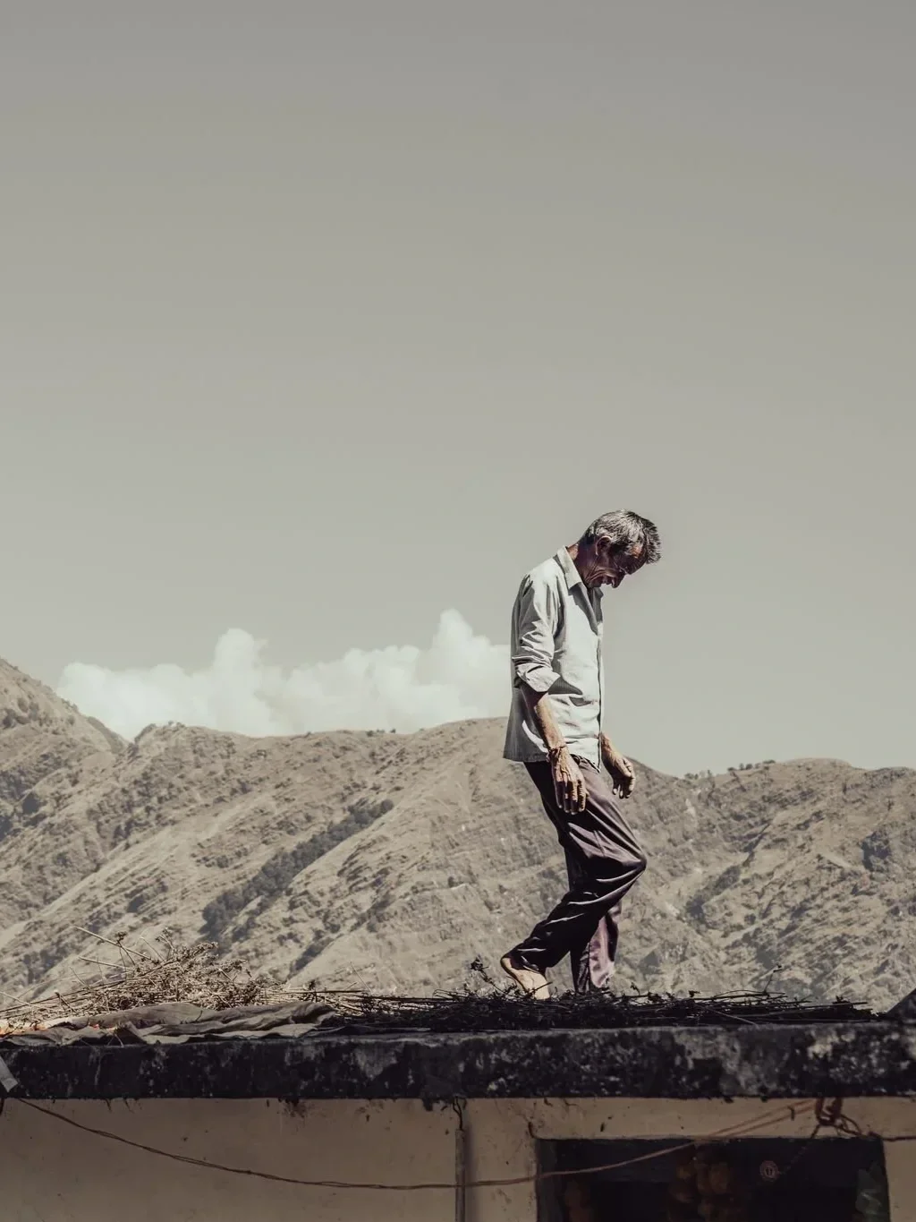 A man with gray hair and a beard standing on a rooftop with mountainous terrain in the background. He appears to be looking down or in deep thought.
