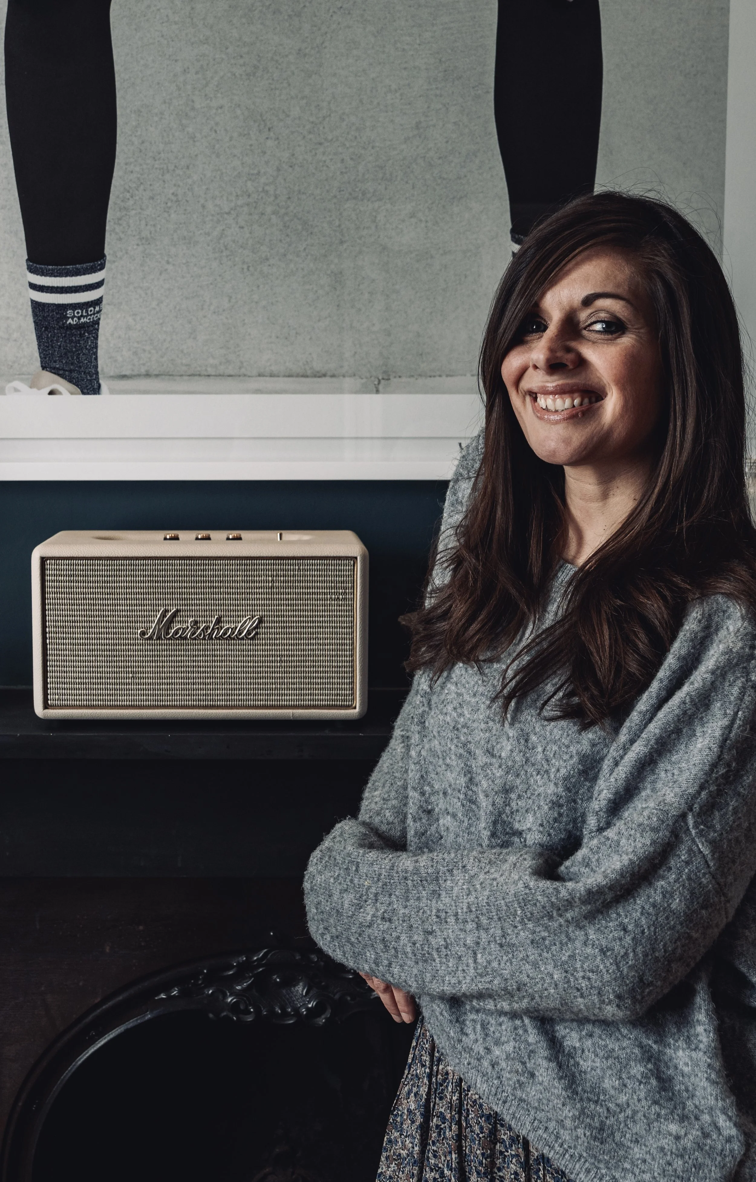 A woman in a gray sweater standing next to a beige Marshall speaker, with a poster of a person in sportswear partially visible behind her.