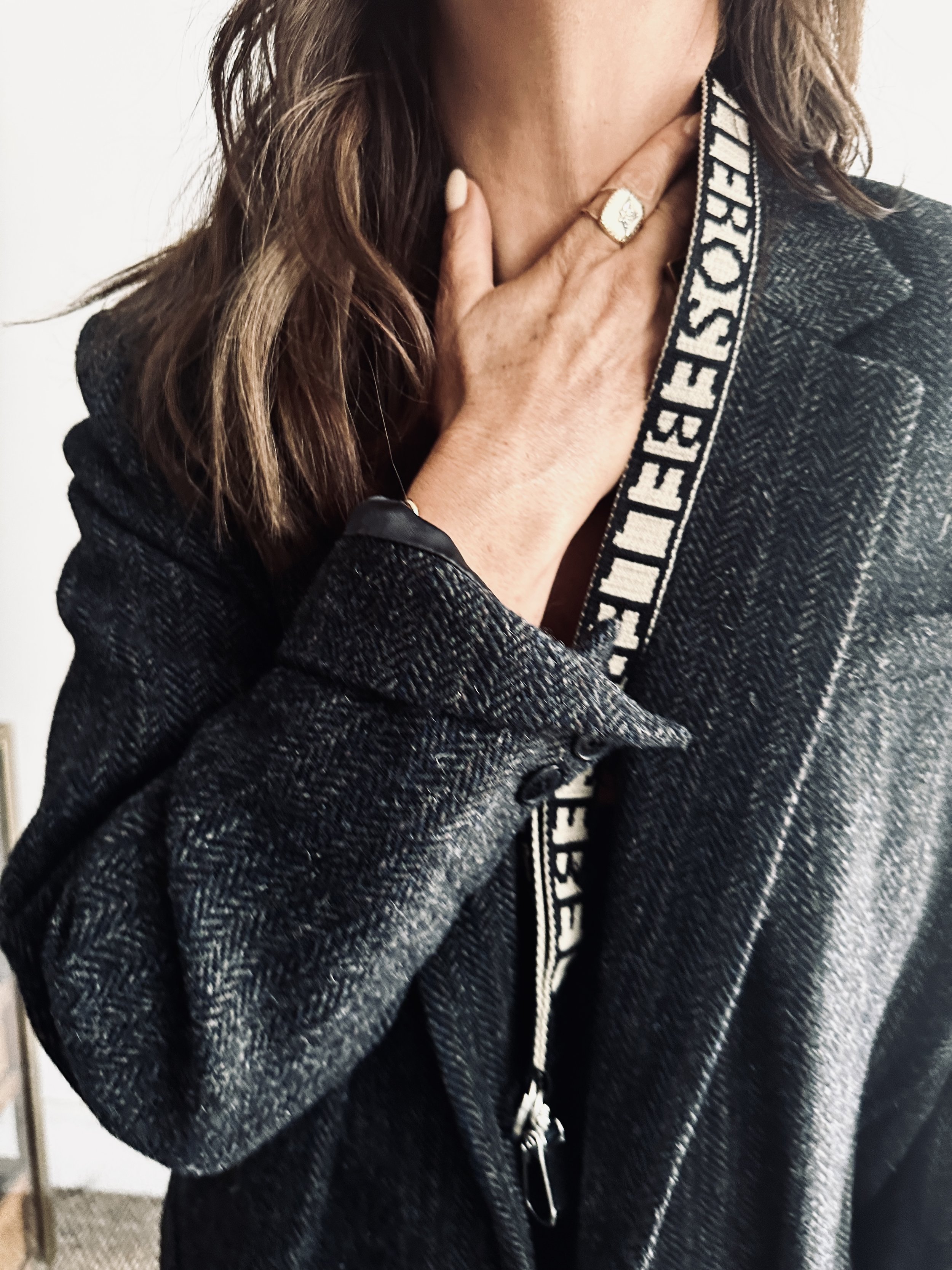 Close-up of a woman wearing a black and white patterned jacket, with her hand on her neck showing a gold ring, and part of a black and white strap with the words "FEED YOUR HUNGER" visible.