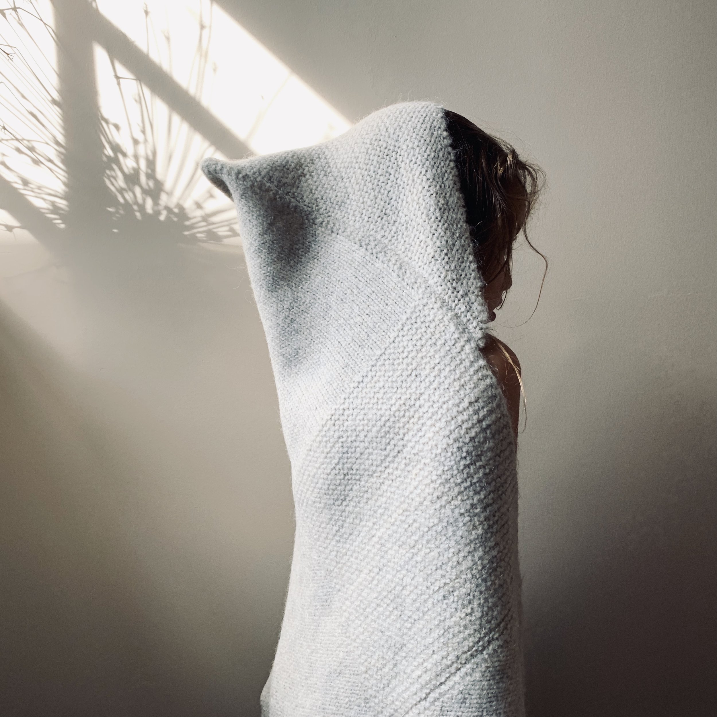 A young child wearing a beige hat and scarf, sitting on a wooden surface, playing with a small stuffed animal and socks, with a plain wall in the background.