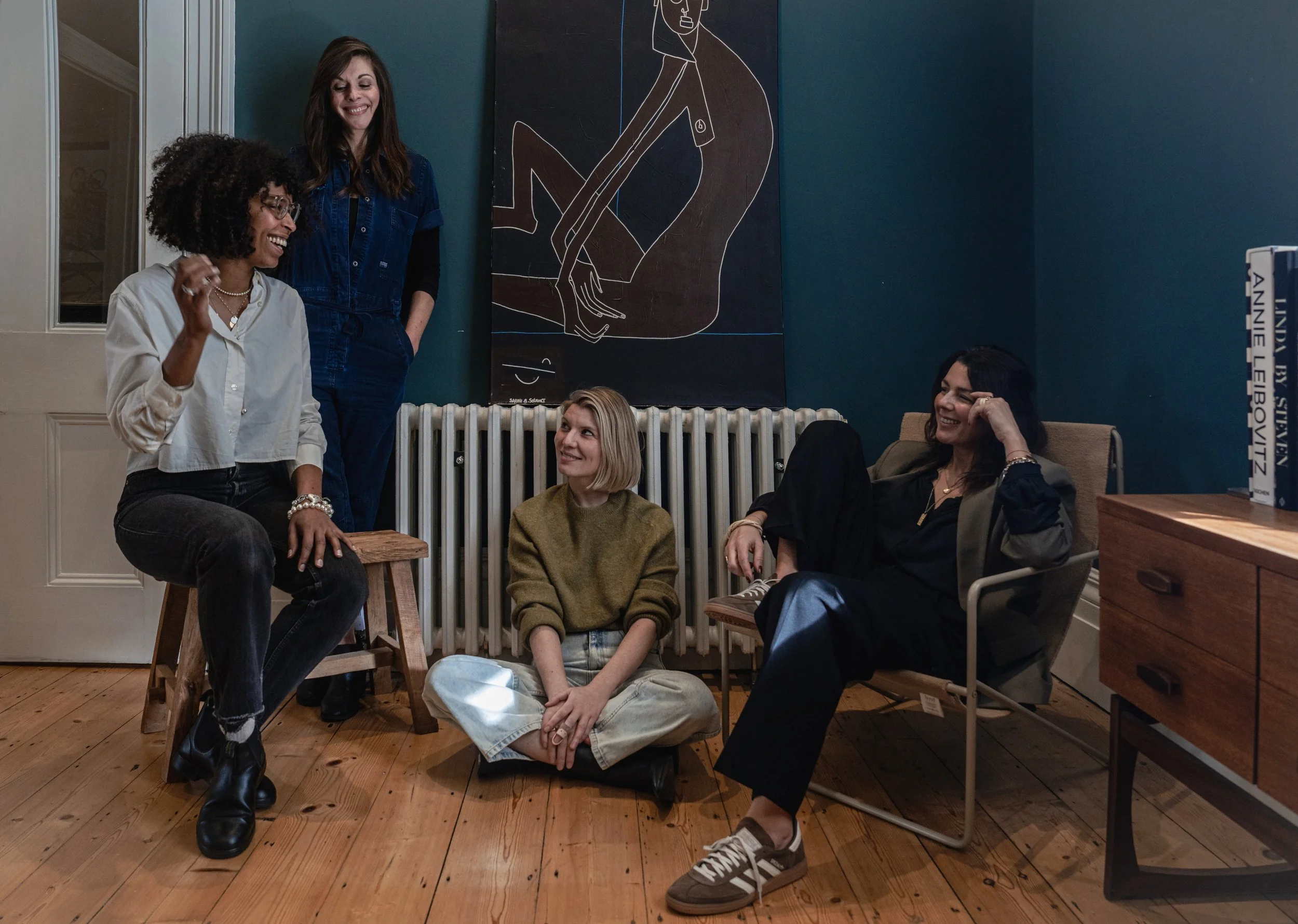 Five women in a cozy room with wooden floors, sitting and standing, engaging in conversation and laughter. A radiator and a dark blue wall with abstract artwork are visible in the background.