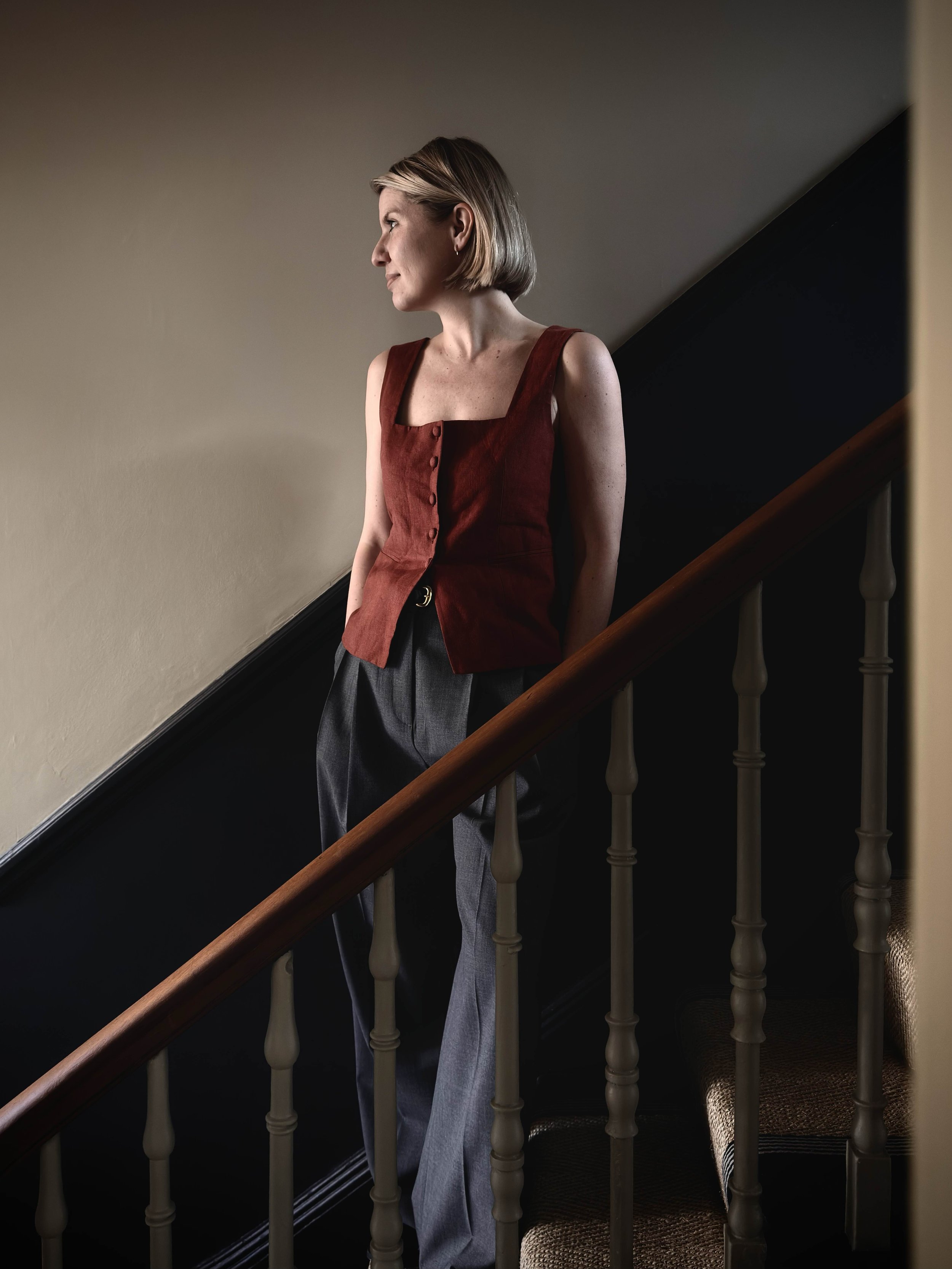 A woman with short blonde hair wears a red sleeveless top and gray trousers while standing on a staircase, looking off to the side.
