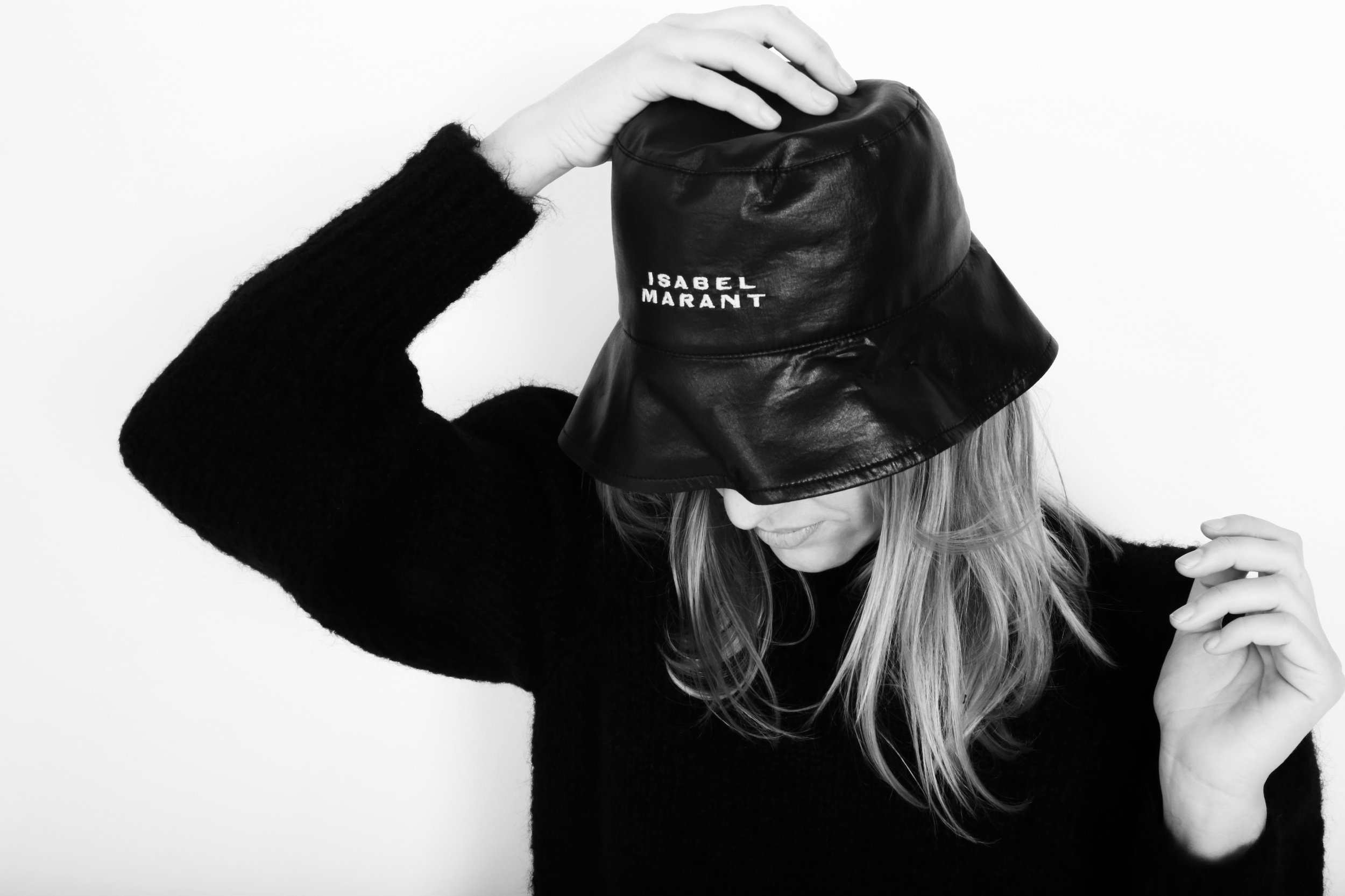 A woman in a black sweater wearing a black leather bucket hat with white text reading 'Isabel Marant', tilting her head downward and adjusting her hat.