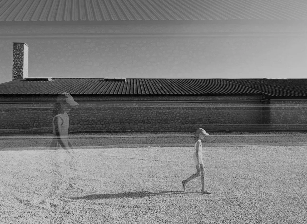 Black and white photo of a person walking on a gravel surface, with a mirrored reflection of the person appearing in a surreal, distorted manner against a building and sky background.