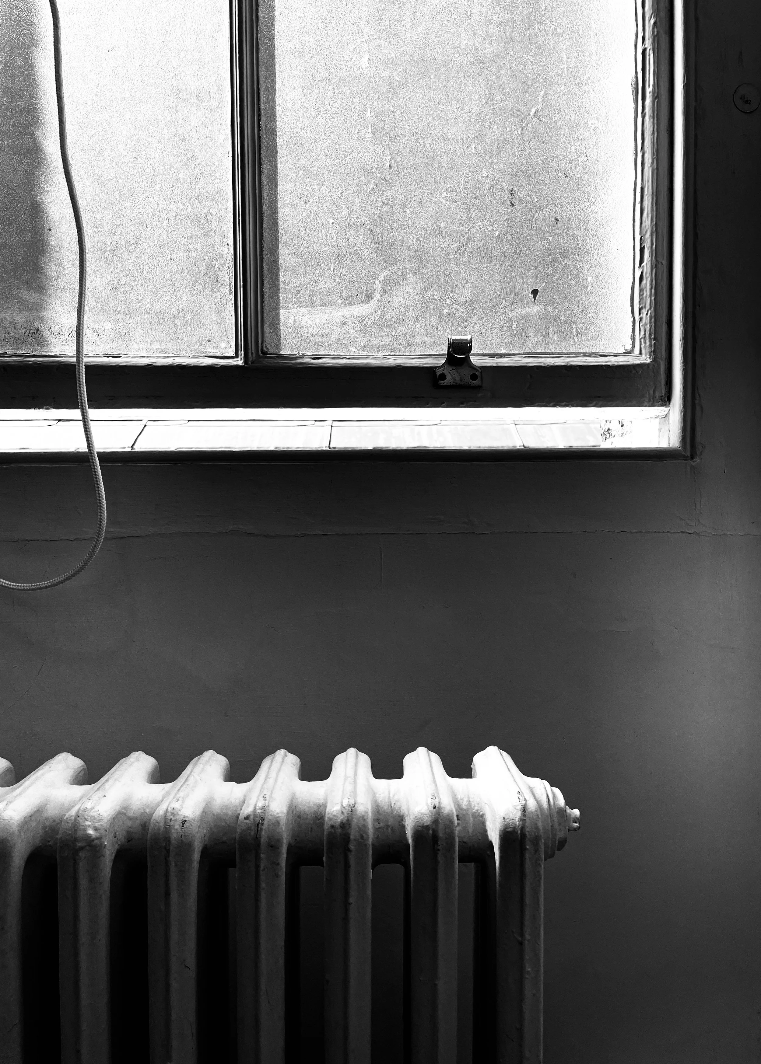 Black and white photo of a window with frosted glass and a latch, above a radiator, with a pull cord hanging beside it.