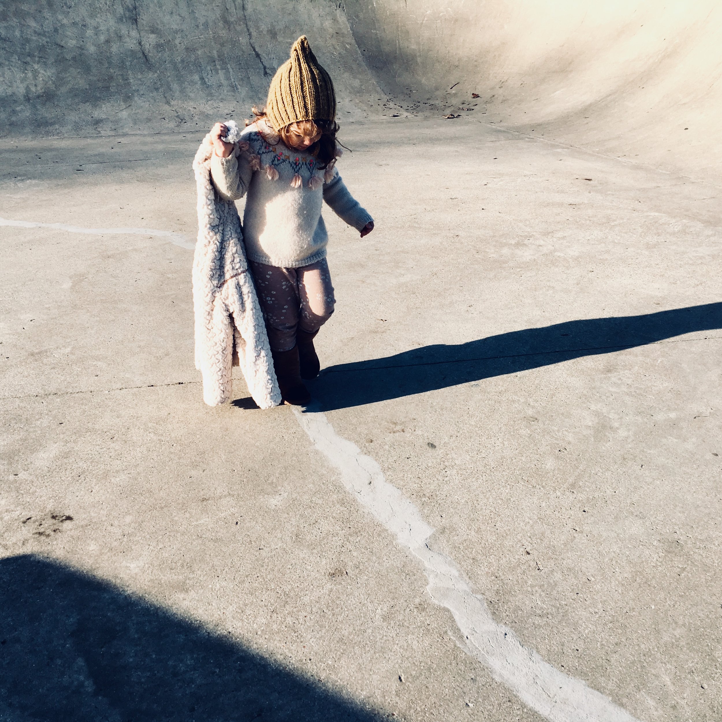 A young girl dressed in warm clothing, including a brown knit hat, sweater, and leggings, standing on a concrete surface with a chalk line tracing her foot.