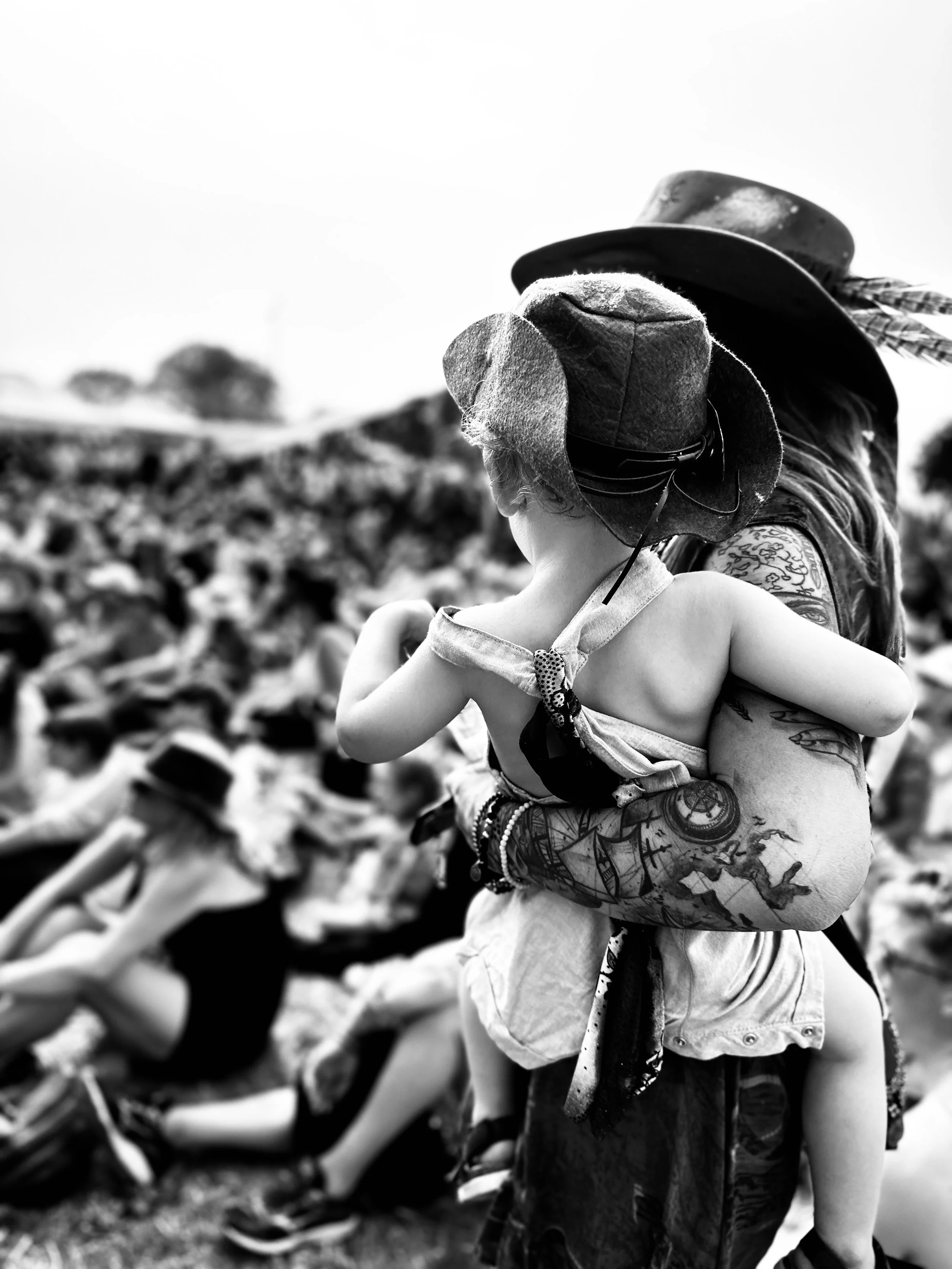 A woman with tattooed arms holding a young child dressed in summer clothes and a hat at an outdoor event with a large crowd in the background.