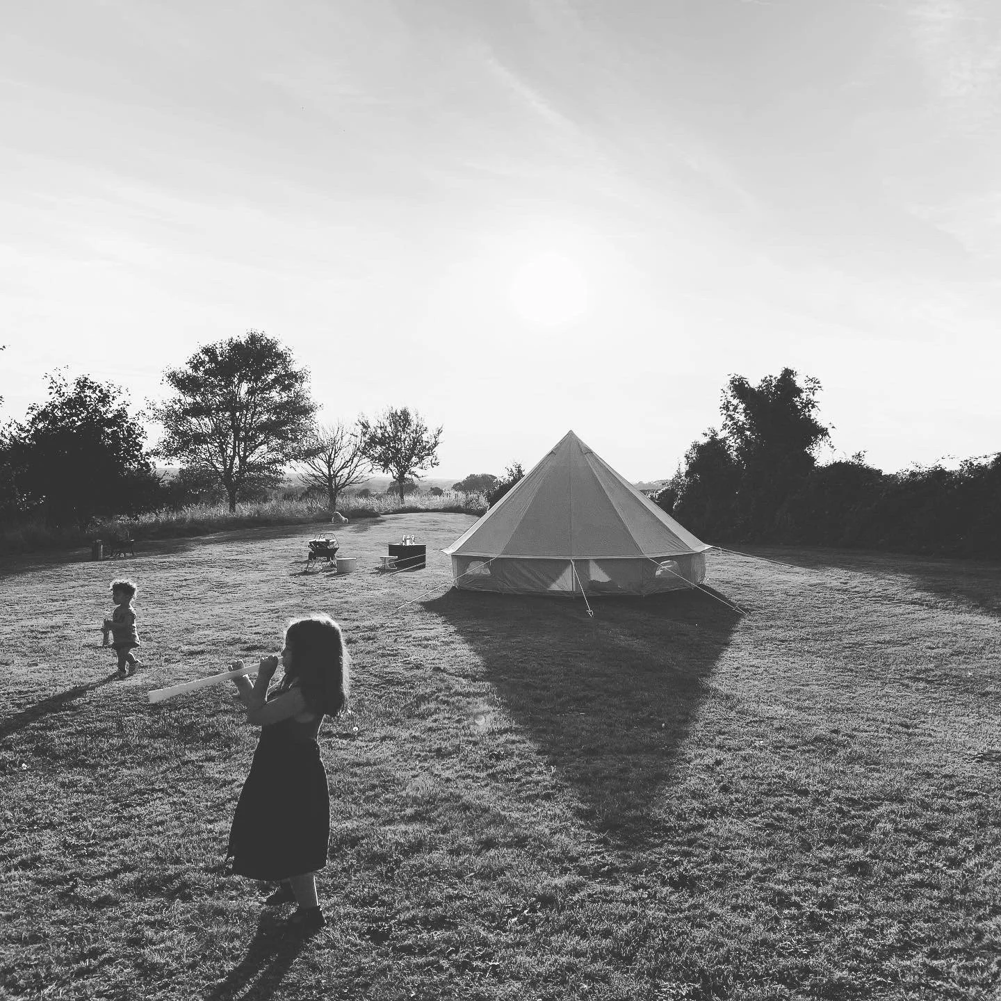 Children playing outside near a tent on a grassy field with trees in the background and the sun setting or rising.