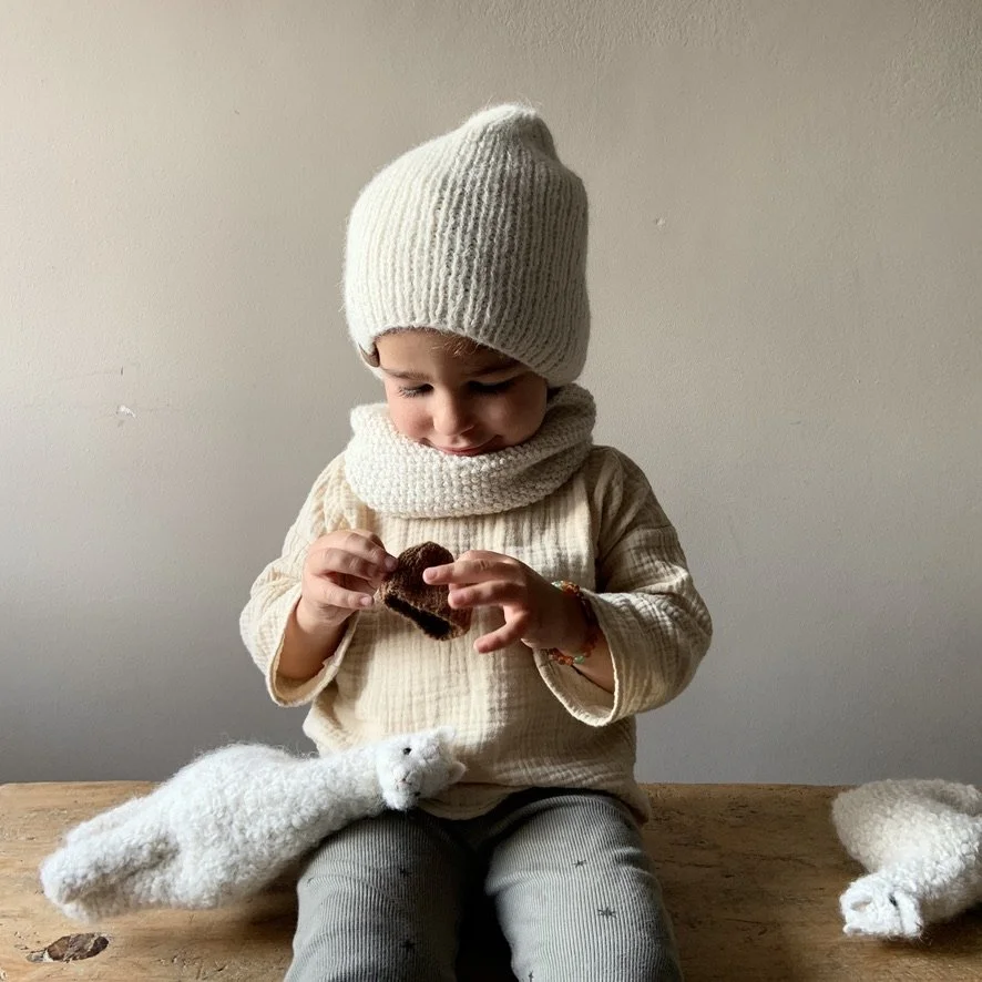 A young child dressed in a beige outfit, knit hat, and scarf, sitting at a wooden table. The child is examining a small dark object while a plush white animal toy, possibly a polar bear, sits on their lap.