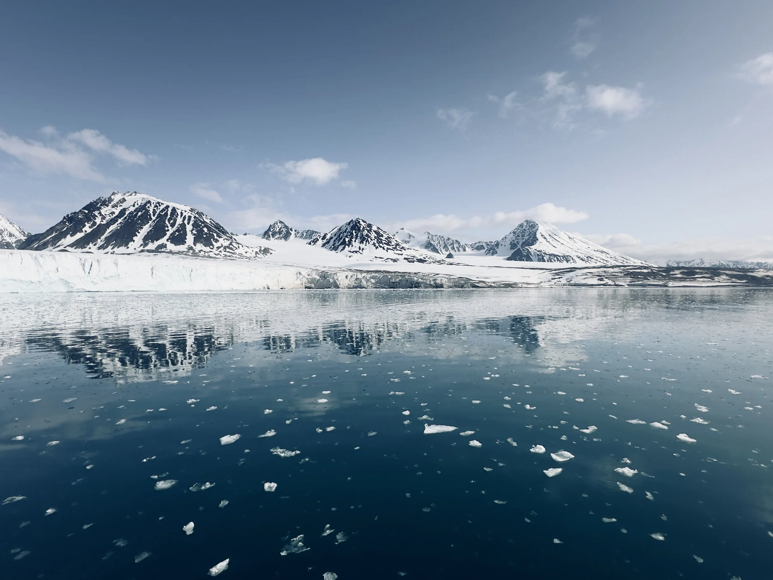 Snow-capped mountains reflecting in icy water with floating ice pieces, under a partly cloudy sky.