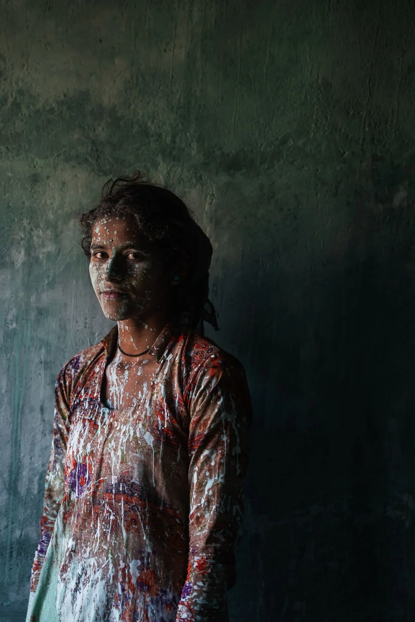 A woman with short dark hair is splattered with white paint, standing in front of a textured dark wall. She is wearing a multicolored patterned garment and has a neutral expression.