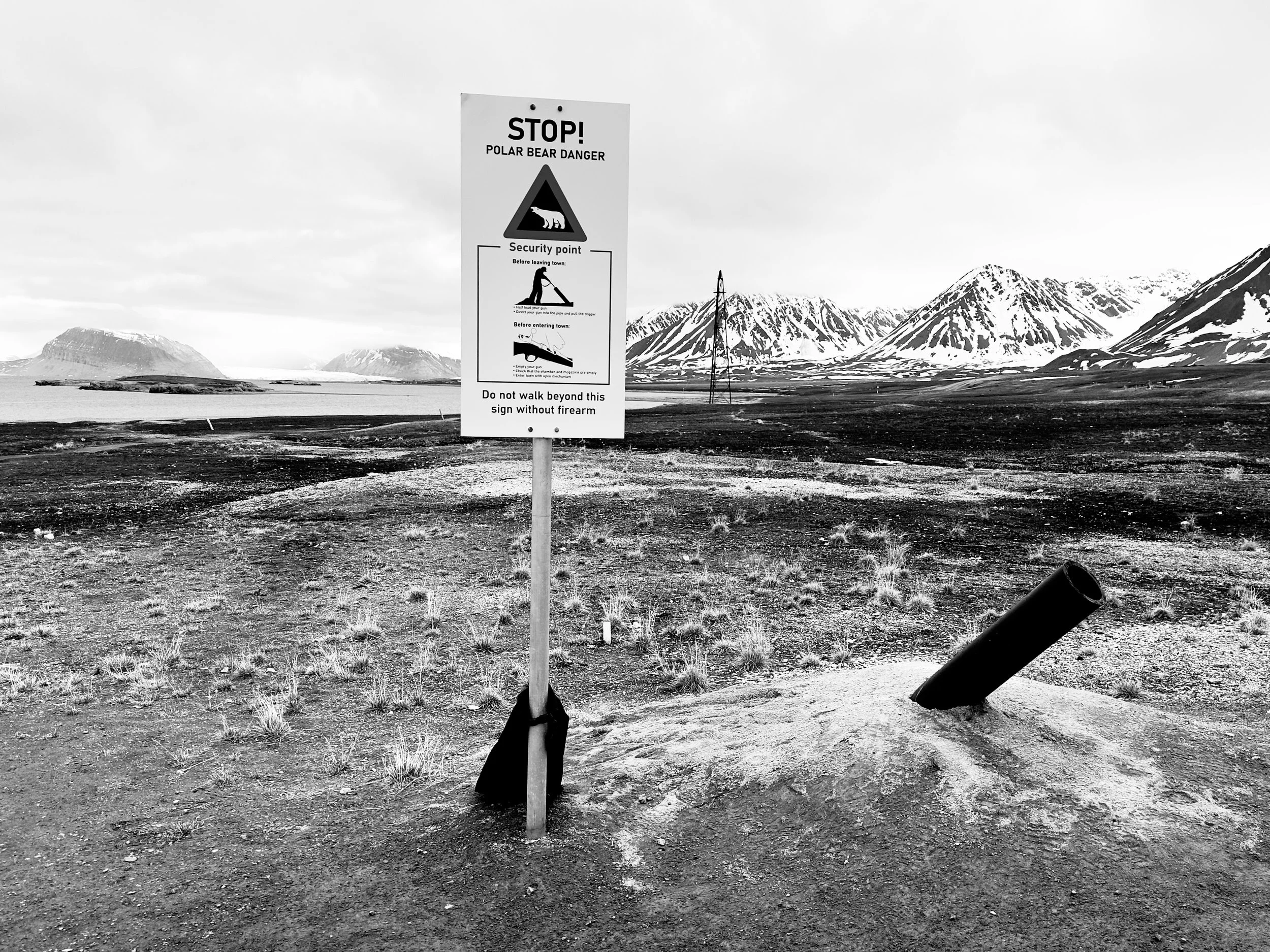 A black and white photo of a sign warning about polar bear danger, with instructions for security and firearm rules, in a barren landscape with snow-capped mountains in the background.
