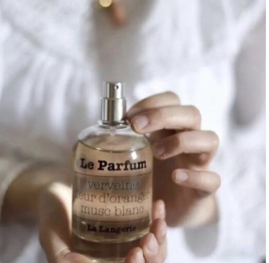 Person holding a clear perfume bottle labeled 'Le Parfum' with scent notes of white musk, orange blossom, and neroli.