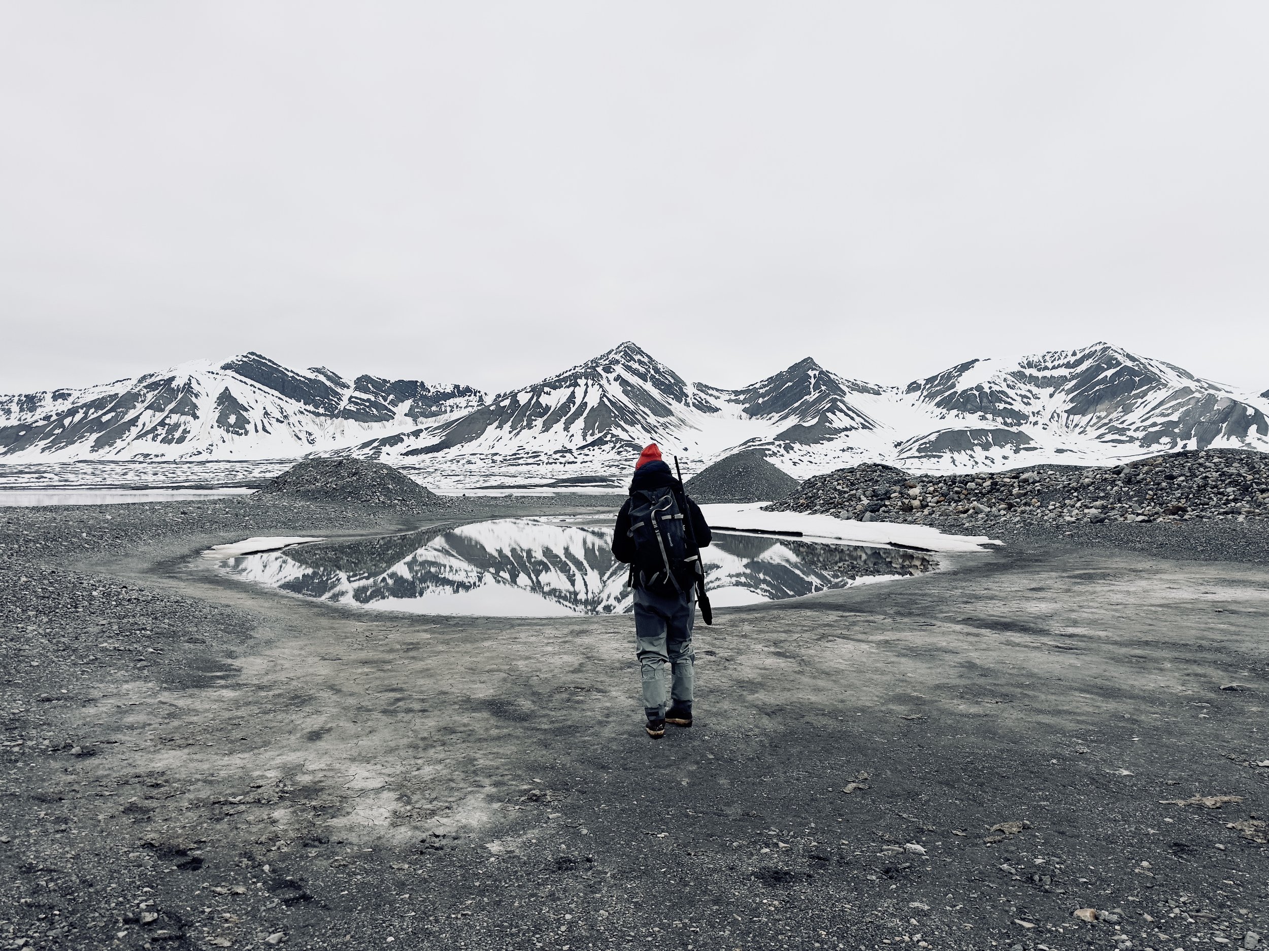 Hiker with a backpack and stick walks towards a reflective body of water with snow-covered mountains in the background.