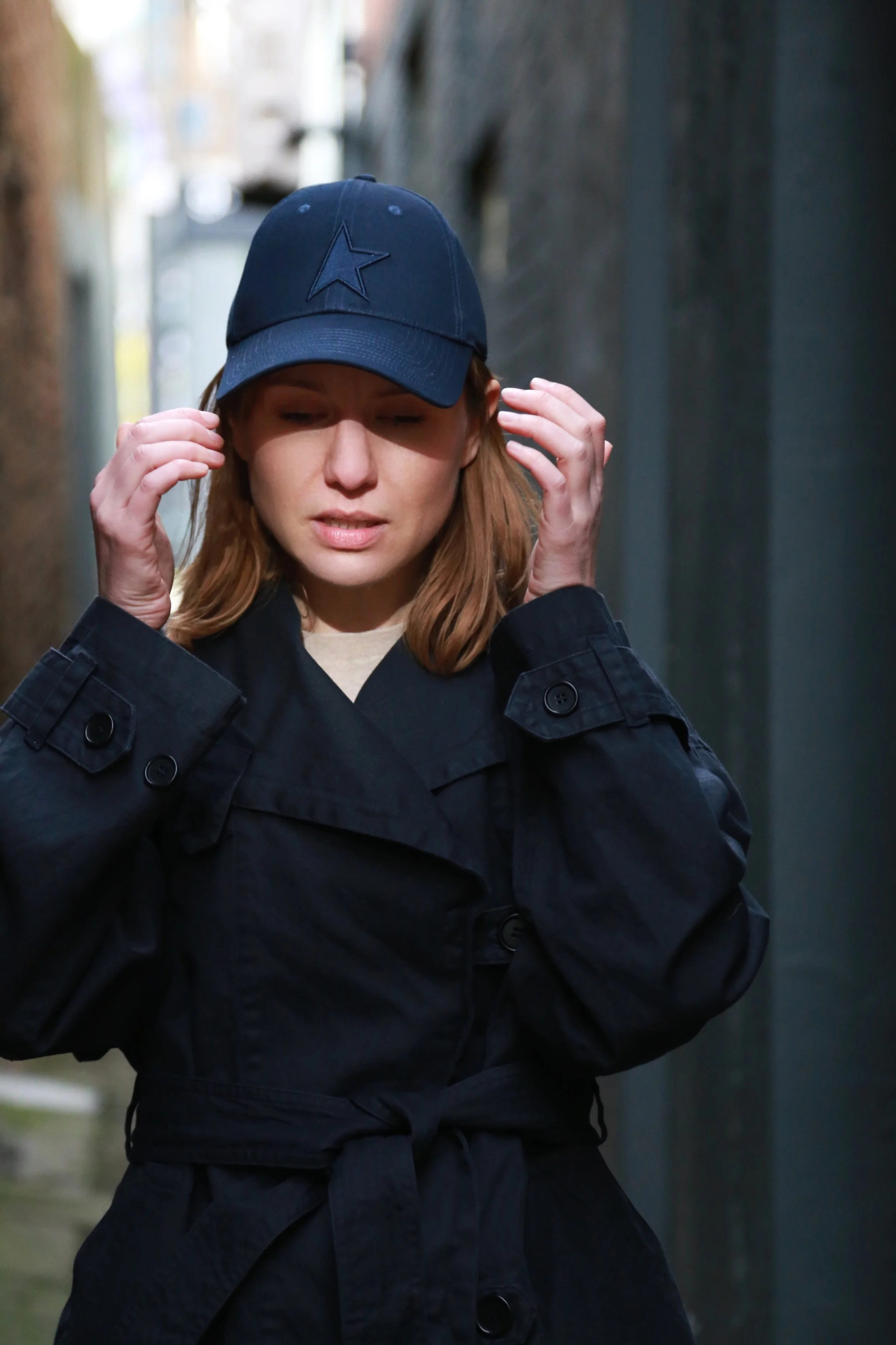 A woman dressed in a black trench coat and wearing a dark blue baseball cap with a star logo, standing in an alleyway, looking thoughtful with her eyes closed and hands near her head.