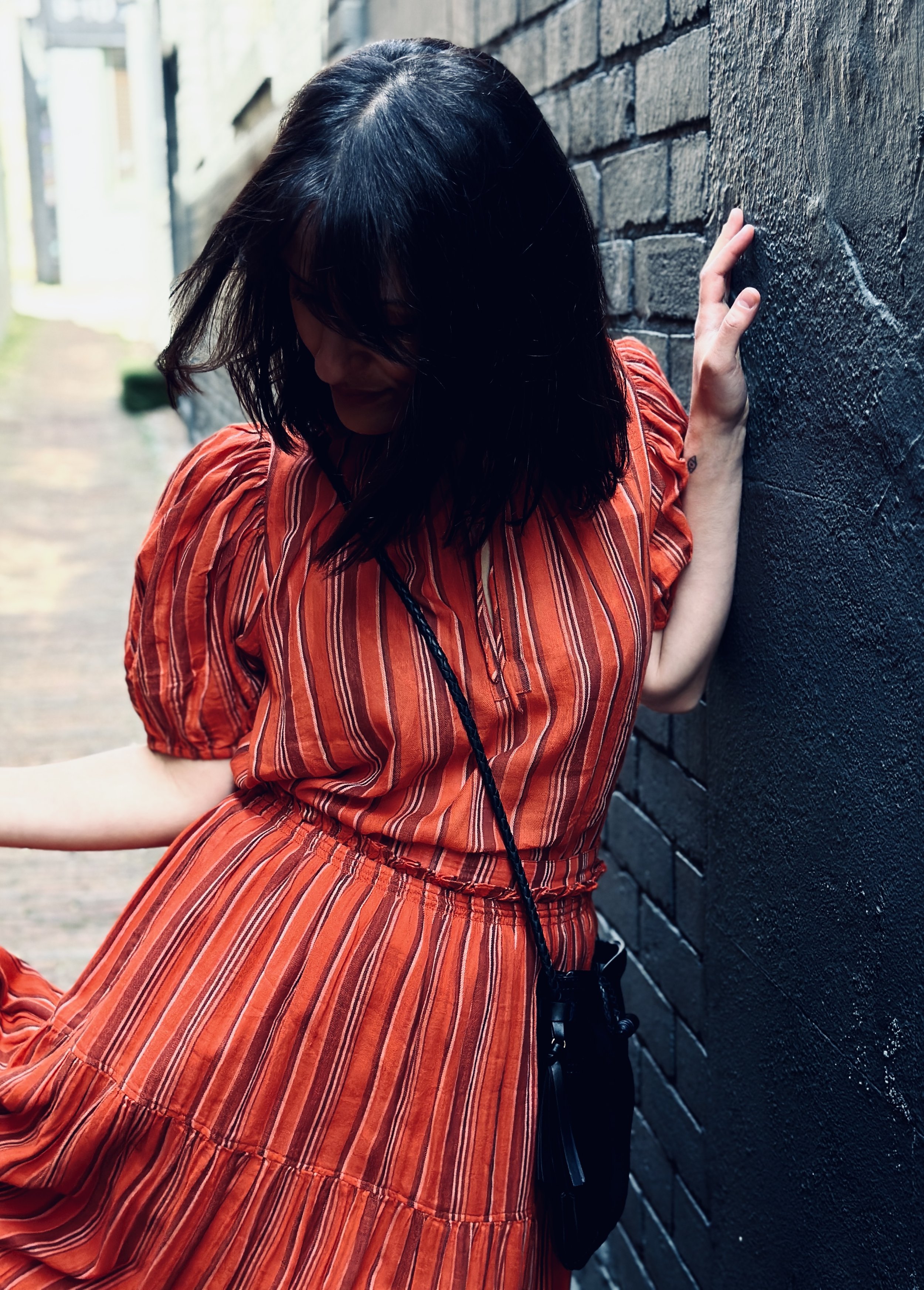 A woman with black hair wearing a red striped dress, leaning against a black brick wall with her head bowed and eyes closed.