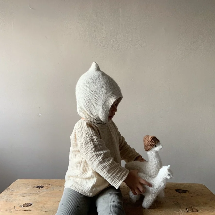 A child wearing a knit hooded sweater playing with a stuffed animal dog with a brown knit cap, sitting at a wooden table against a plain beige wall.