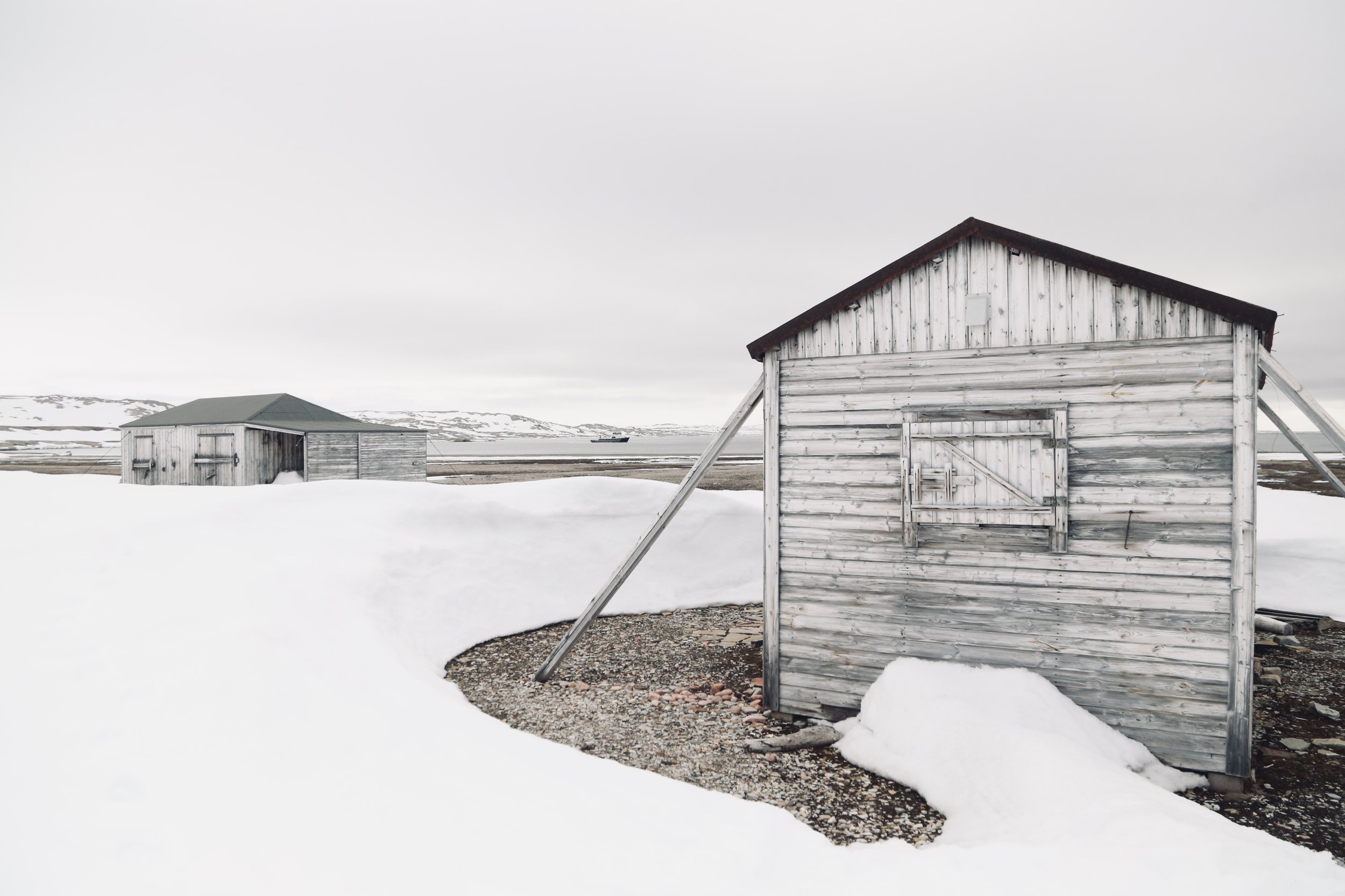 Two weathered wooden sheds in a snowy landscape with distant mountains and cloudy sky.