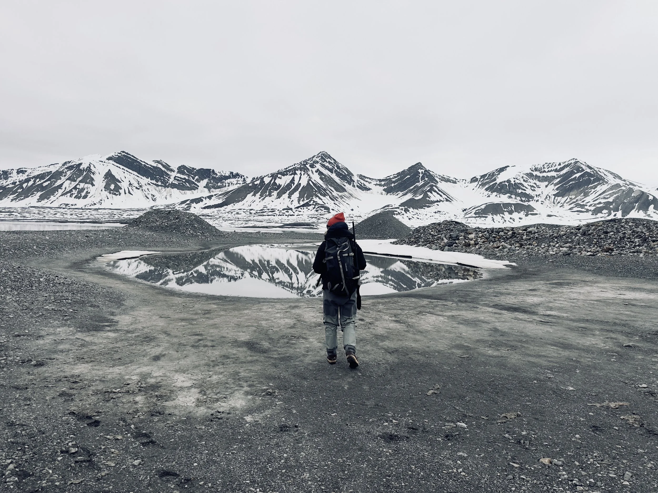 A person with a backpack hiking in a barren, rocky area near a reflective body of water, with snow-covered mountains in the background under an overcast sky.