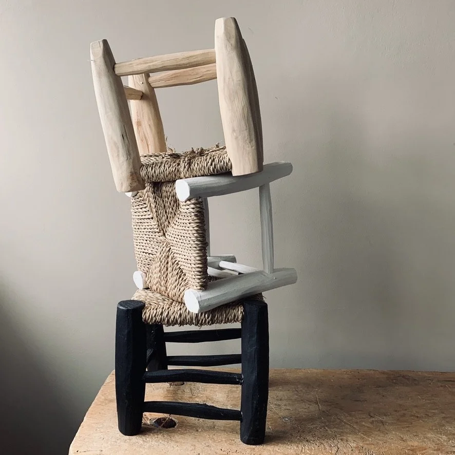 Stacked wooden chairs with woven seats, arranged to resemble a sculpture or abstract art piece on a wooden table.