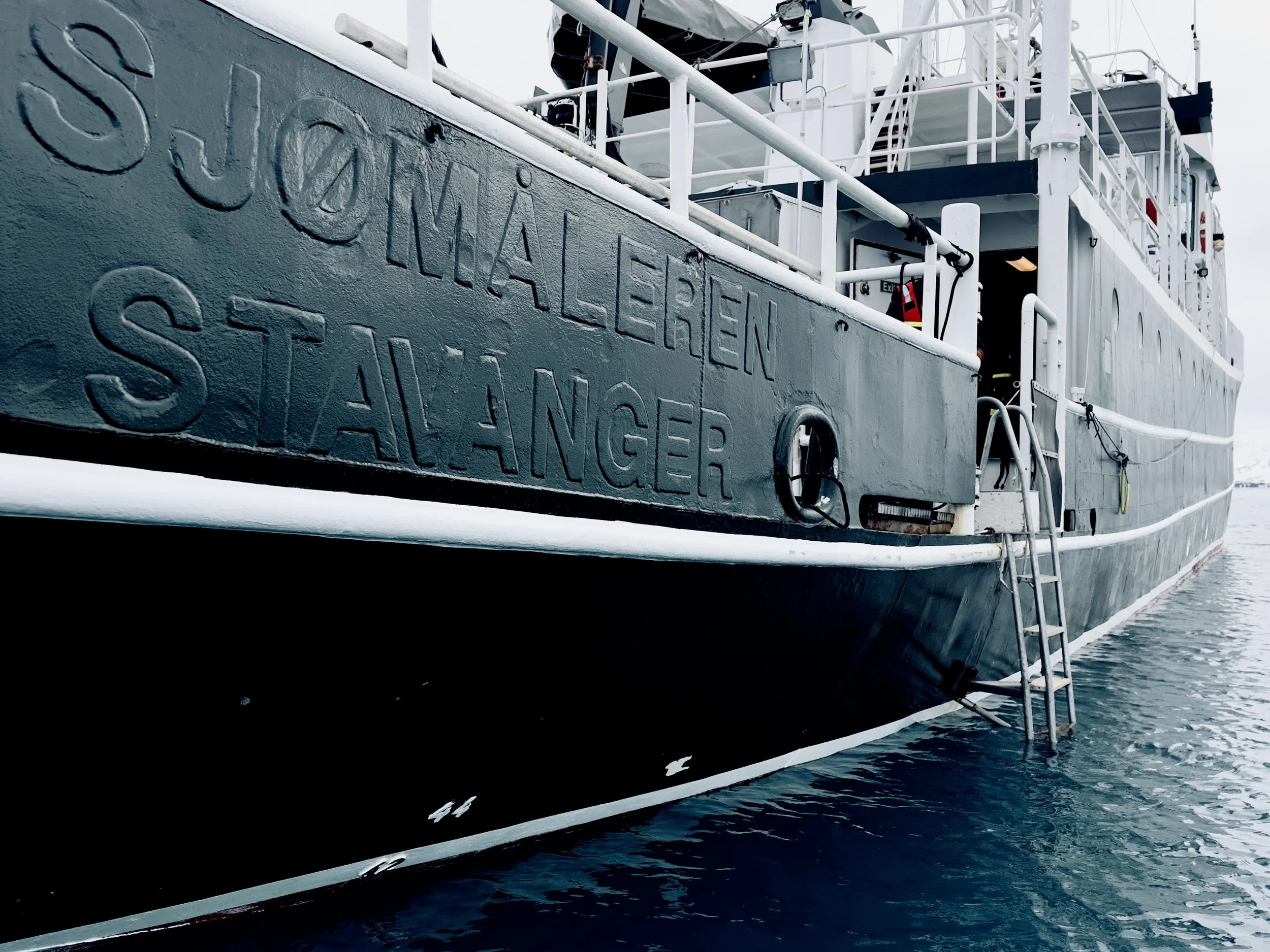 Close-up of the side of a gray boat with the words 'Sjømælerieren Stalanzer' and a ladder hanging over the water.