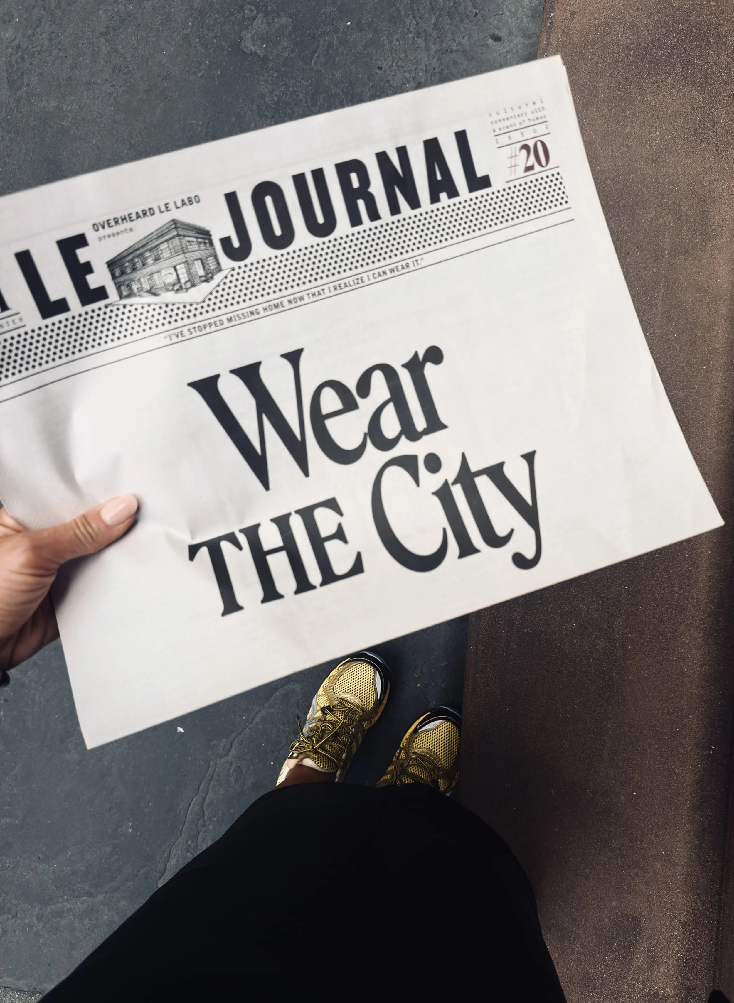 Hand holding a newspaper with "Wear the City" headline, gold trainers on a New York pavement, photographed by Clementine for MUSE