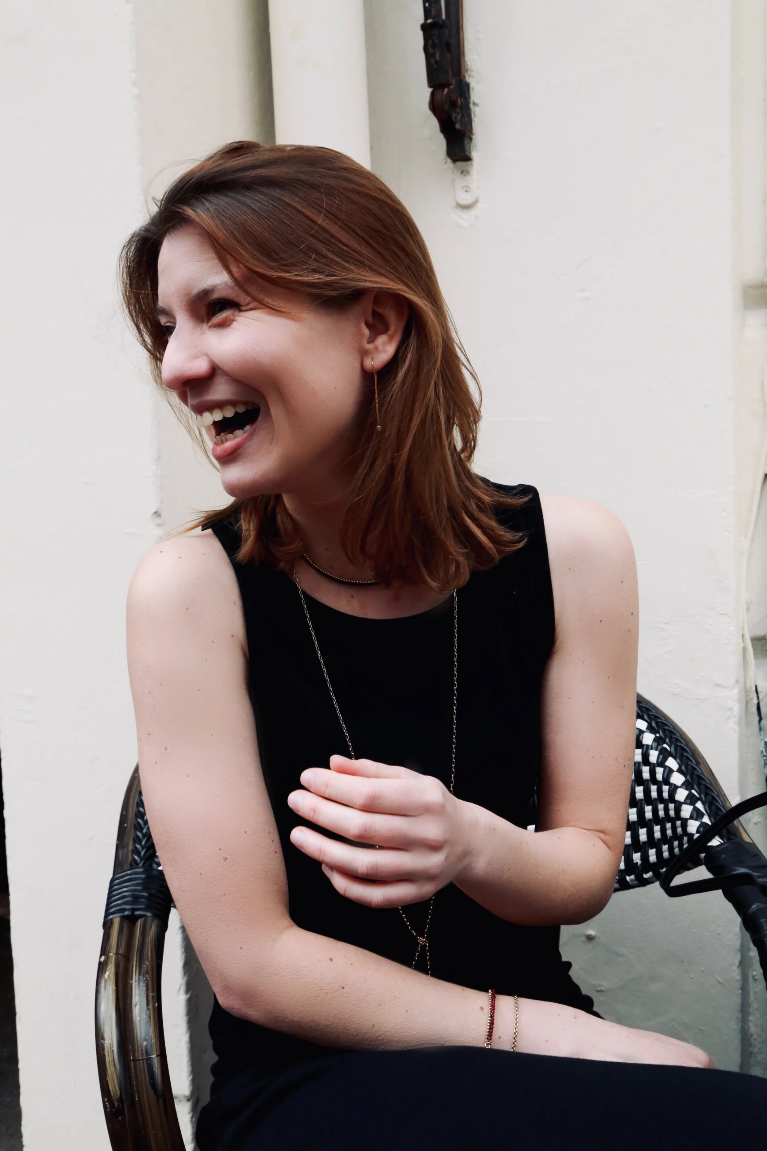 A woman with red hair, wearing a black sleeveless top and jewelry, is smiling and laughing while sitting on a wicker chair indoors.