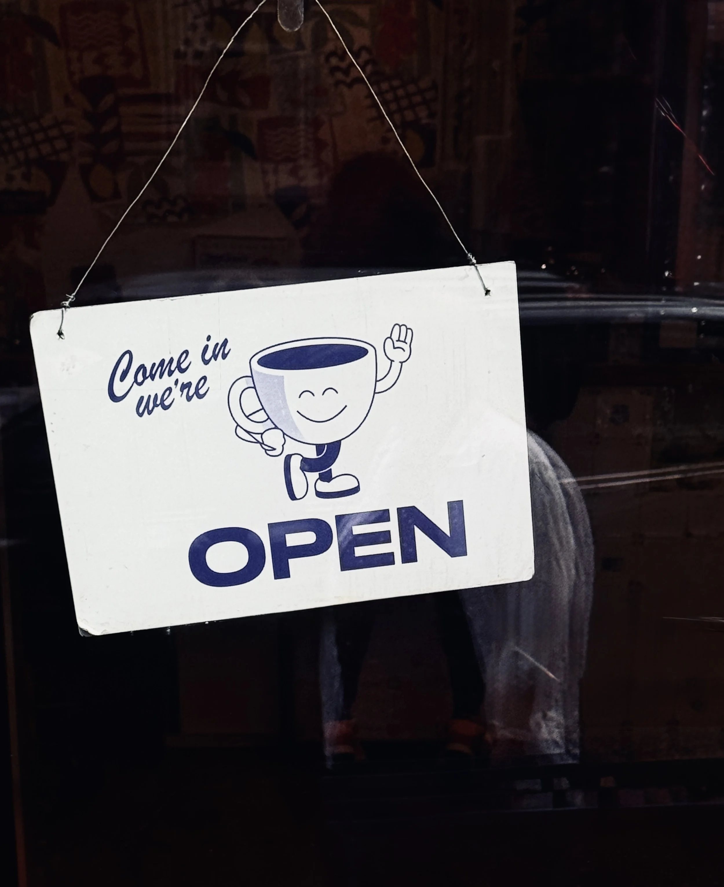 "Come in, we're open" sign with a smiling coffee cup illustration in a New York café door