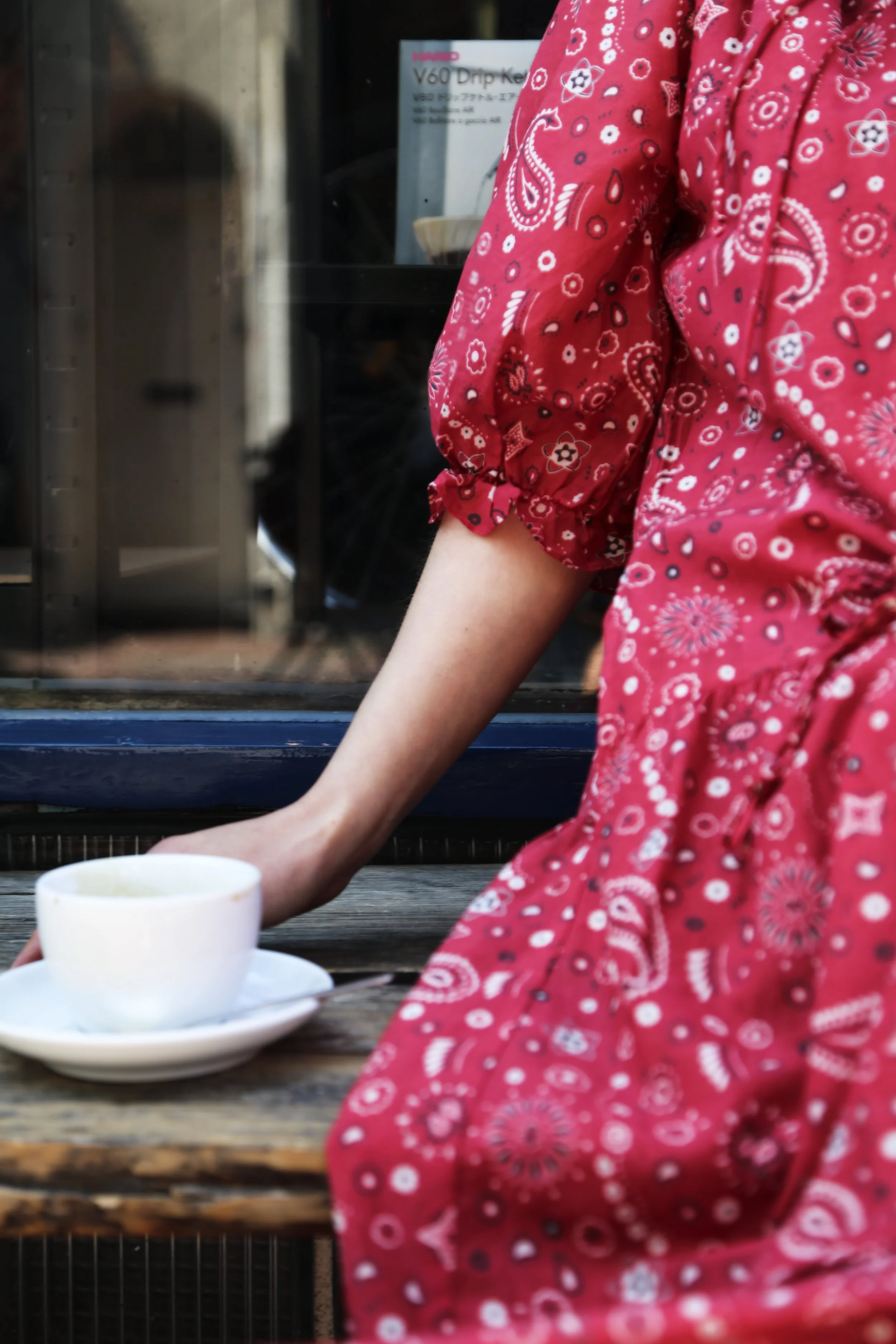 A person wearing a red floral-patterned dress with puffed sleeves, reaching for a white teacup on a saucer placed on a rustic wooden table outside a cafe.