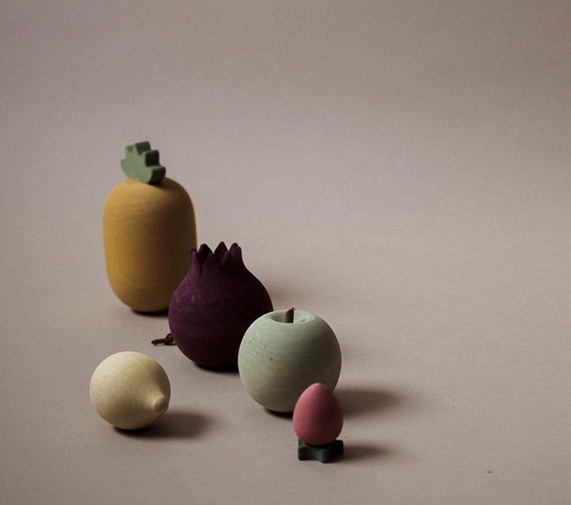 Various colorful wooden fruit decorations including a pineapple, pomegranate, apple, lemon, and a small pink fruit with a bow, arranged on a plain surface.