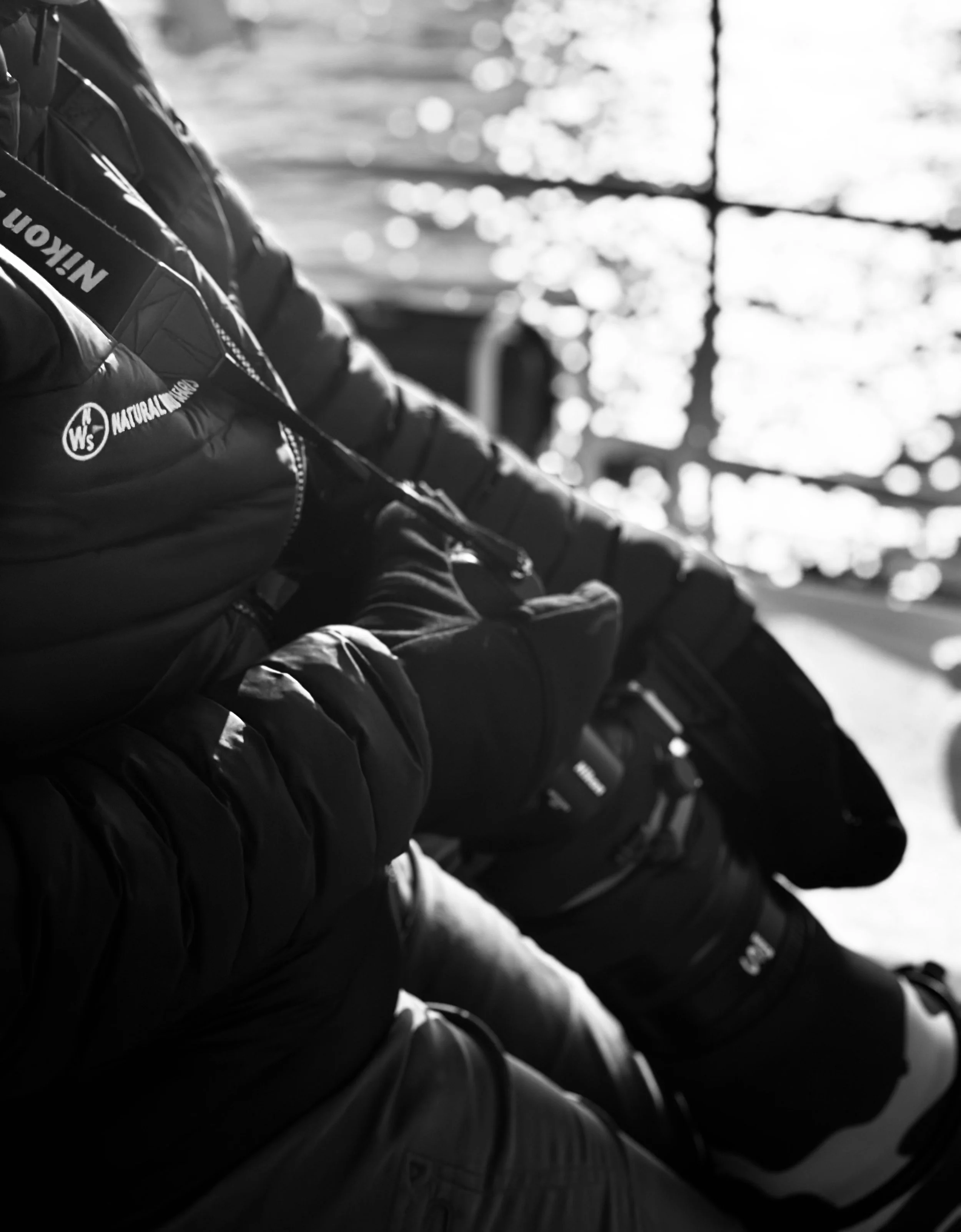 Black-and-white photo of a row of people sitting on a bench, dressed in winter clothes, outdoors with sunlight filtering through trees in the background.