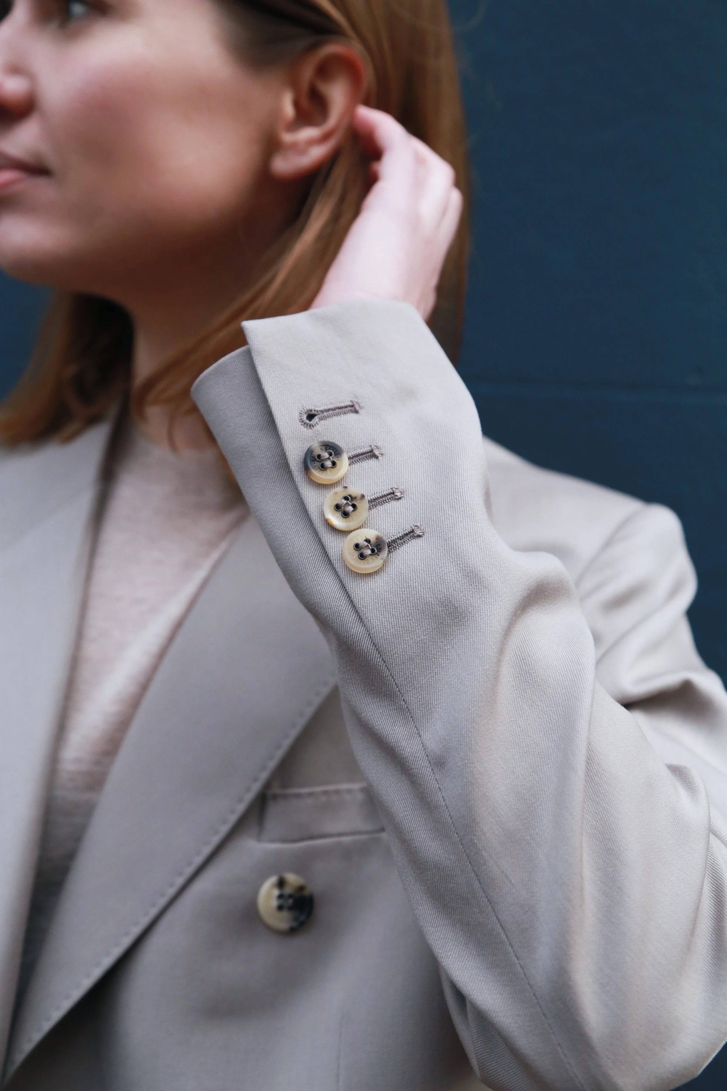 Close-up of a woman's arm wearing a beige blazer with four buttons on the sleeve, adjusting her hair with her hand. The background is dark blue.