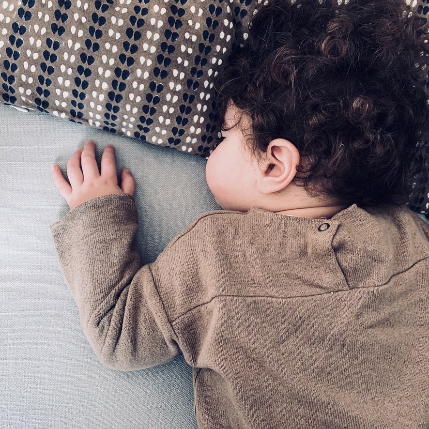 A young child with curly dark hair sleeping on a light-colored surface with a patterned pillow nearby.