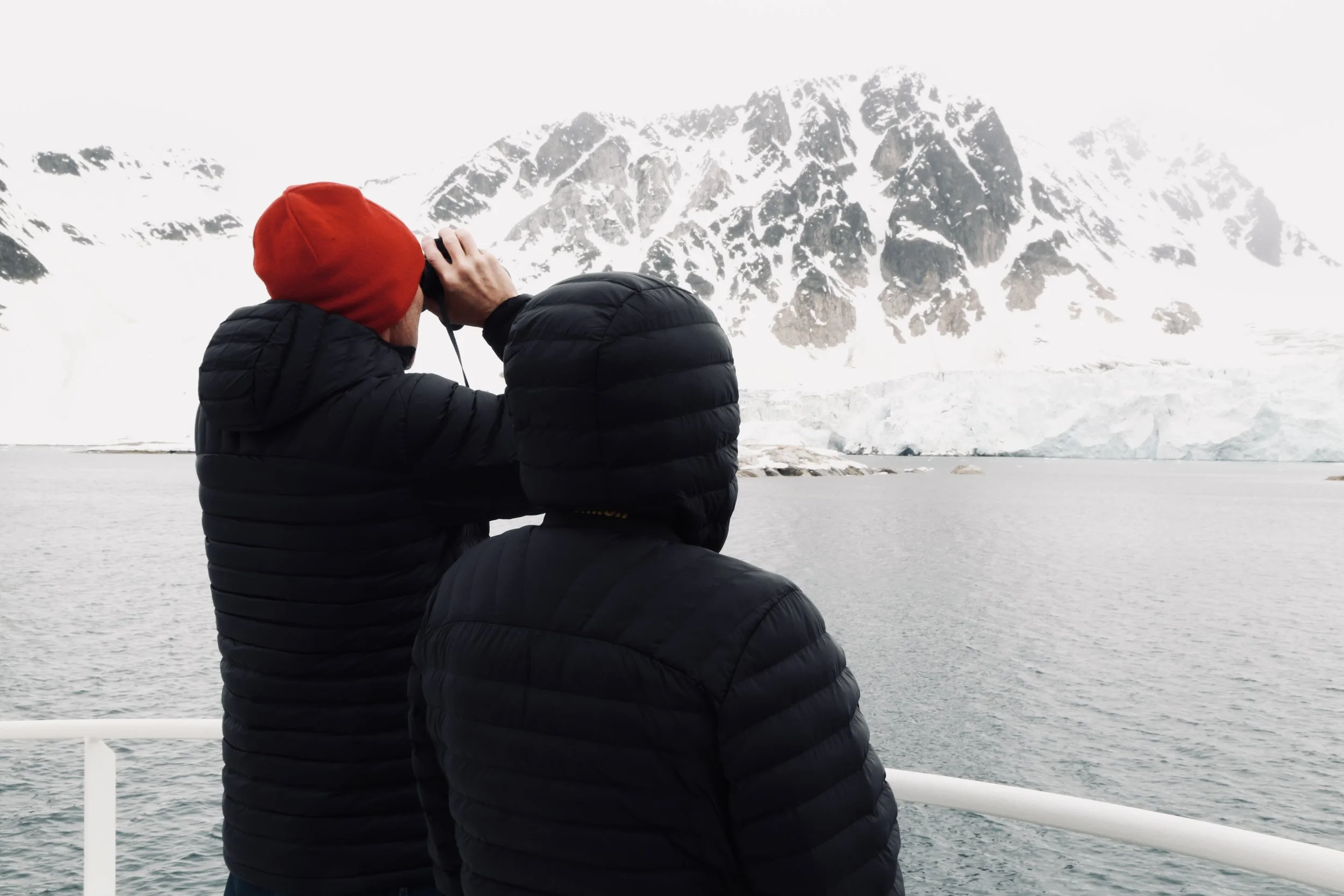 Two people in black jackets and red, black hats observing icy landscape through binoculars on a boat in a cold, snowy environment