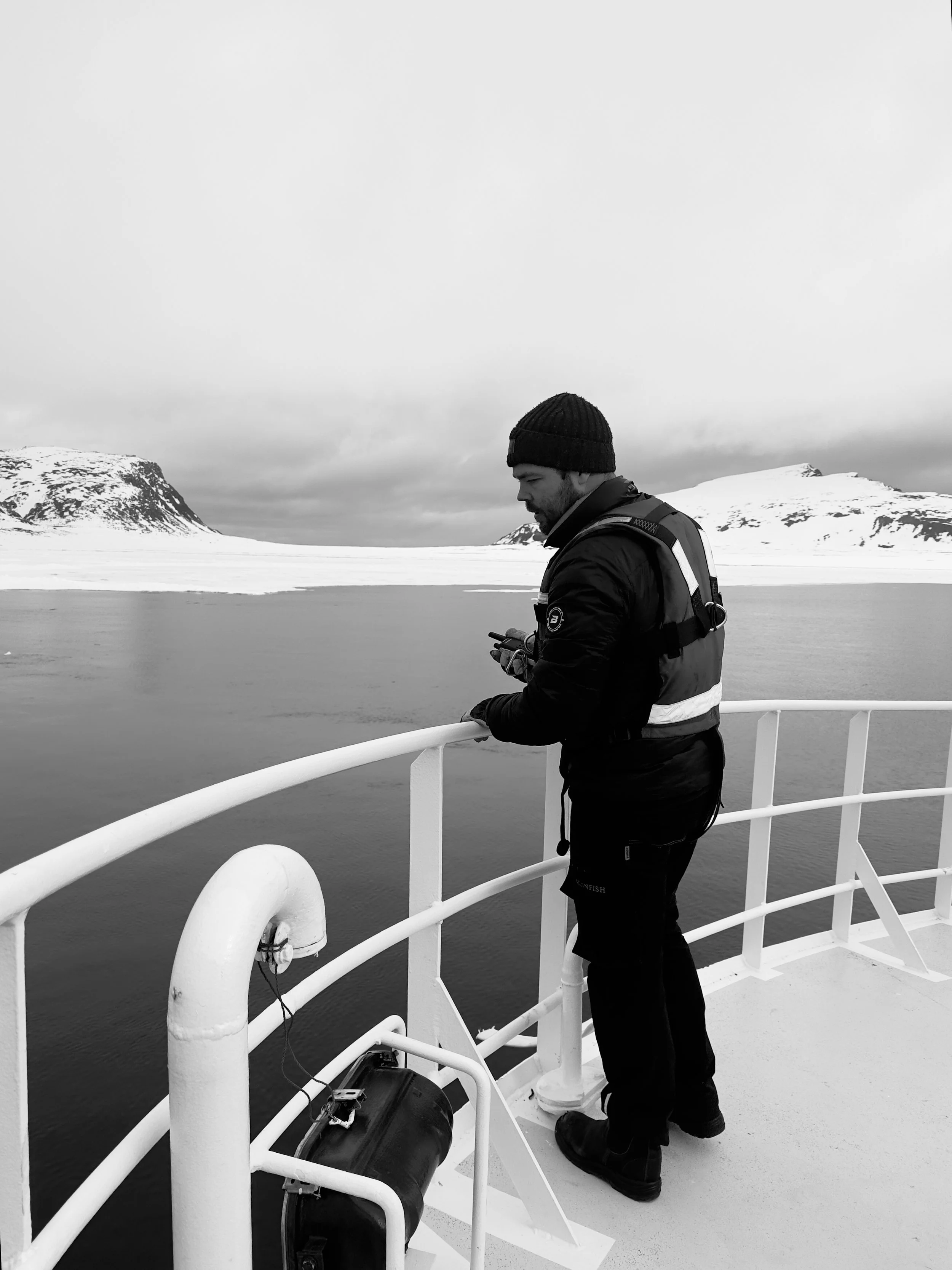 A man wearing a beanie and a jacket standing on a boat's deck, looking at his phone, with snow-covered mountains and water in the background.