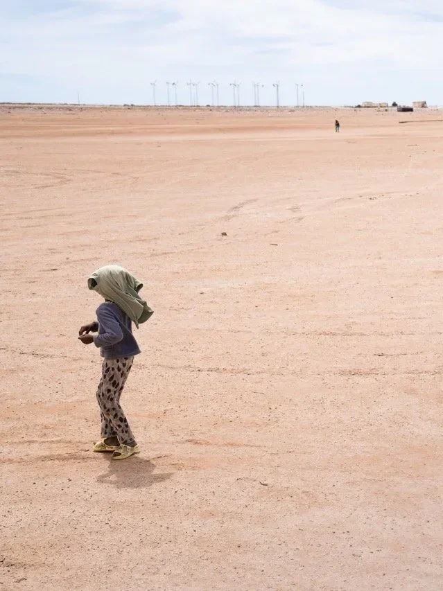 Child wearing a headscarf and patterned pants standing on a vast, empty desert landscape with wind turbines in the background.