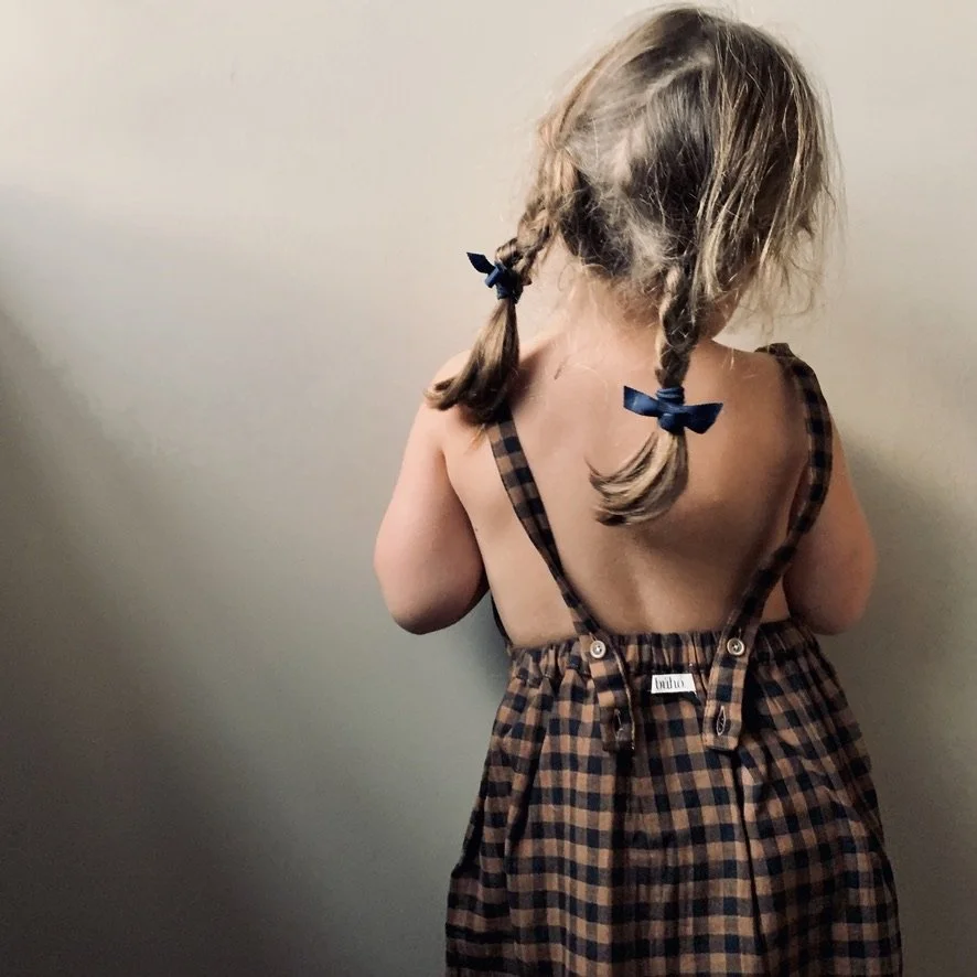 A young girl with blonde hair in pigtails wearing a checkered dress, facing away from the camera, standing against a plain wall.