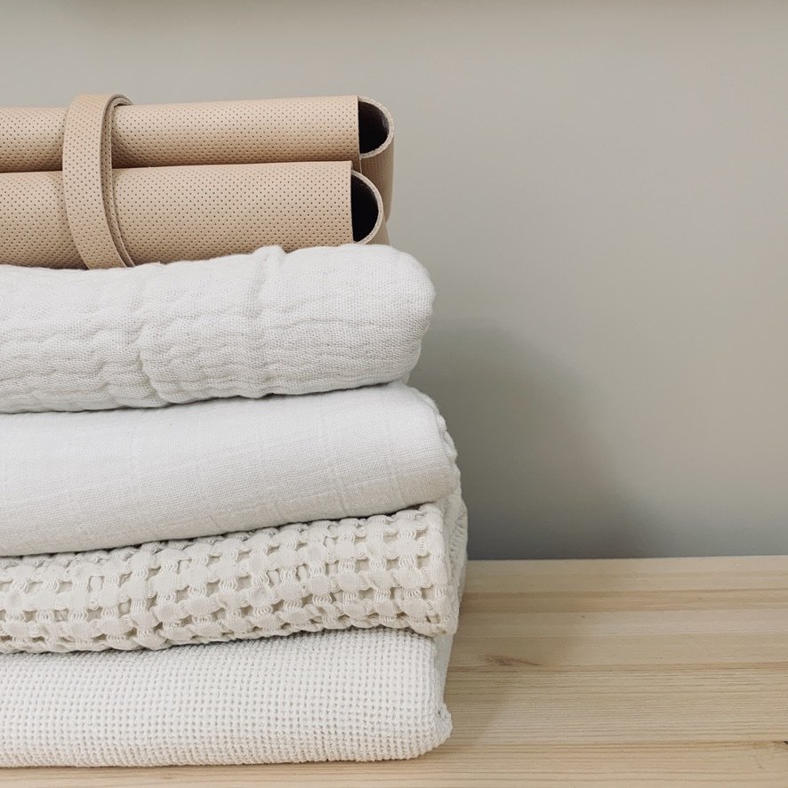 Stack of folded white and beige textiles with rolled up beige foam rollers on top, on a wooden surface against a gray wall.