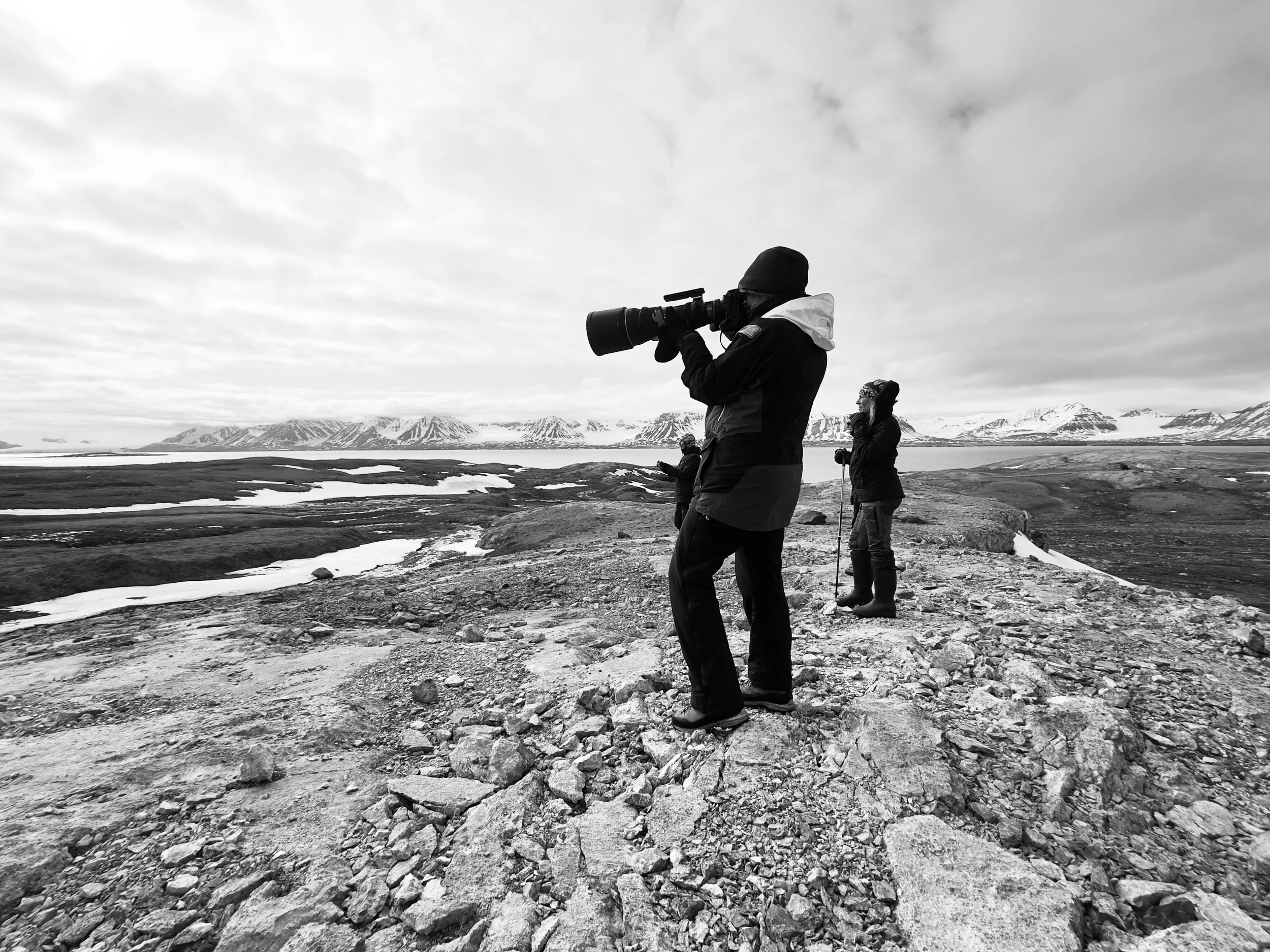 Three people in winter clothing standing on rocky terrain near water, with mountains and snow patches in the background, one person holding a camera with a large telephoto lens.