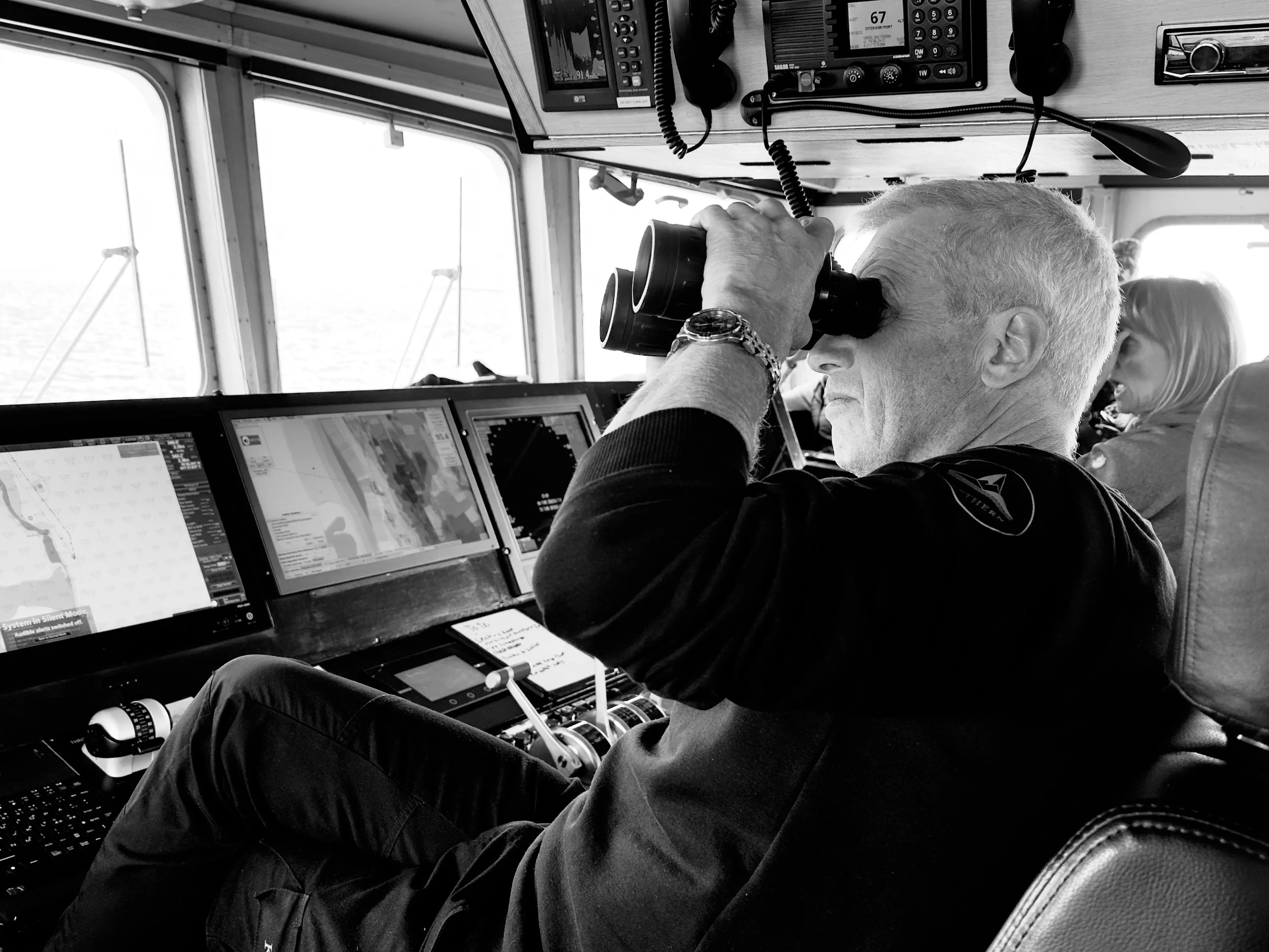 A man with gray hair looking through binoculars in the cockpit of an aircraft, with a woman in the background, computers and navigation equipment visible.