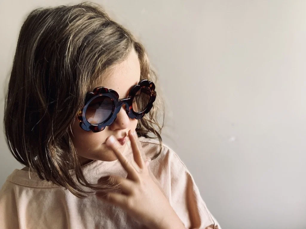 A young girl with shoulder-length brown hair wearing large, dark sunglasses and a beige shirt, holding her fingers close to her lips in a thoughtful pose against a plain light-colored background.