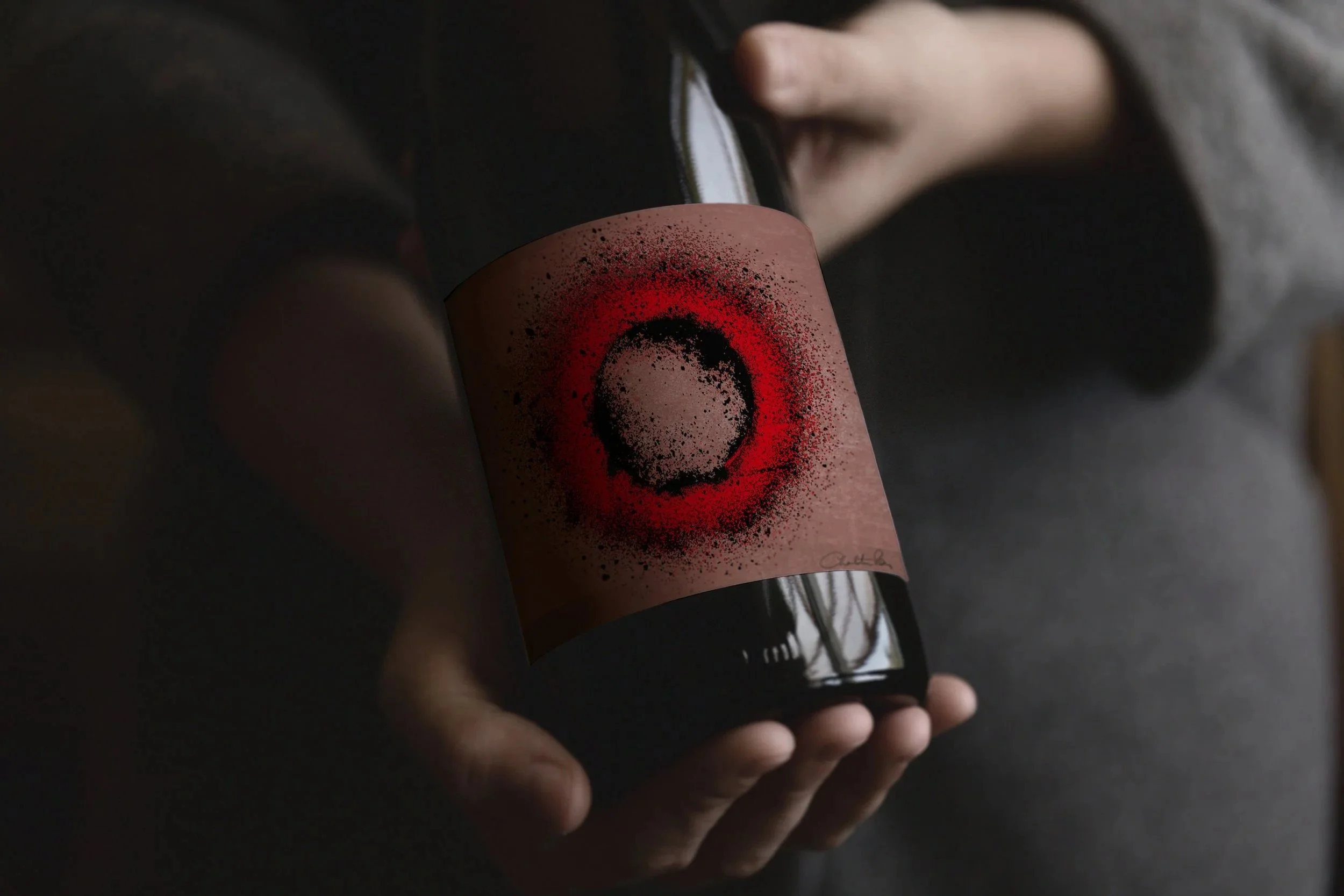 Close-up of a hand holding a dark wine bottle with a pink label featuring black and red concentric circles, against a blurred background.