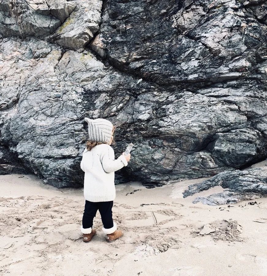 A young child wearing a white knitted sweater, tan boots, and a gray beanie with stripes stands on sandy ground near large dark gray and black rocks, looking at a phone in their hand.