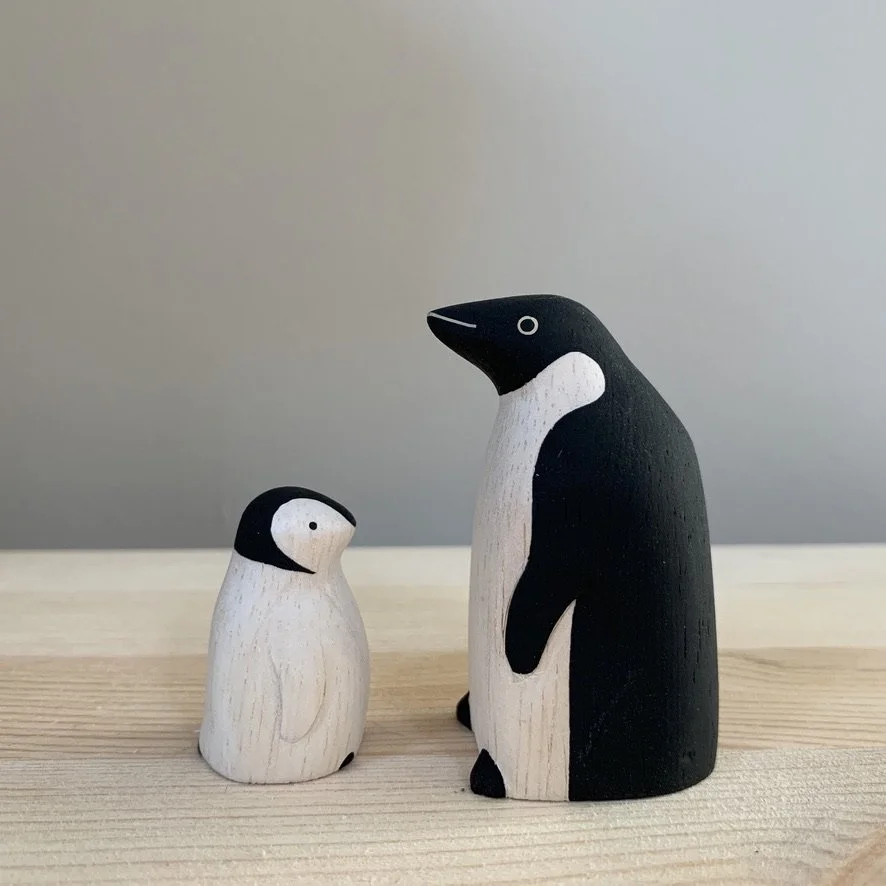 Two painted wooden penguin figures, one large and one small, on a light wooden surface against a gray wall.