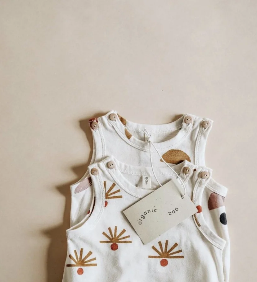 Three white baby tank tops with colorful abstract patterns, stacked on a beige surface, with a tag labeled 'organic zoo'.